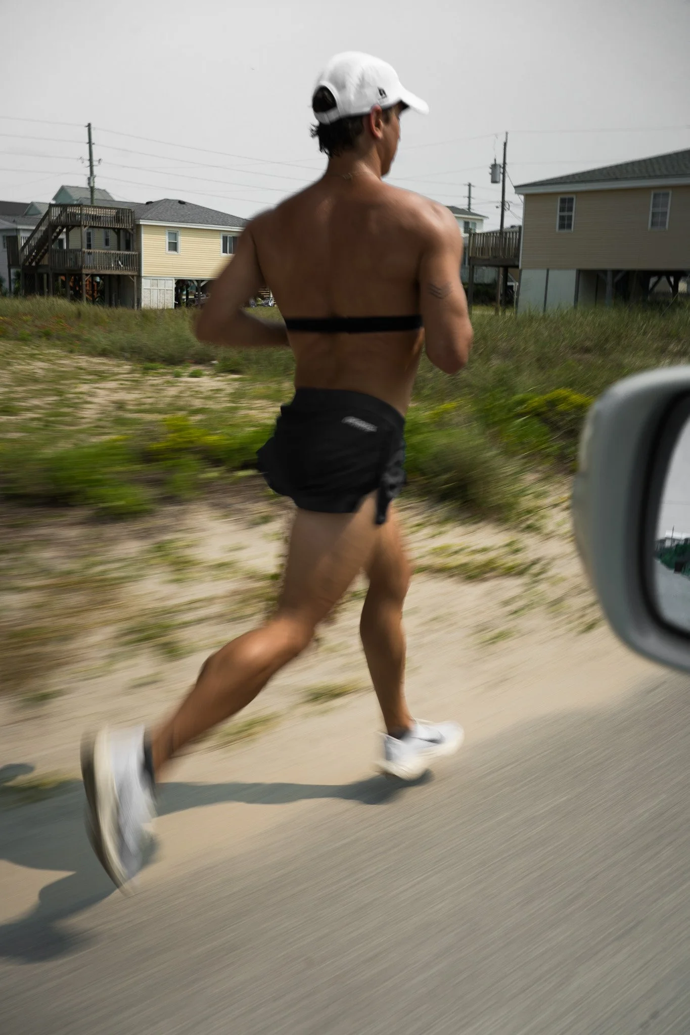 Ian Fisher running on the roadside wearing a white cap, black shorts, and white running shoes, with houses and power lines in the background.