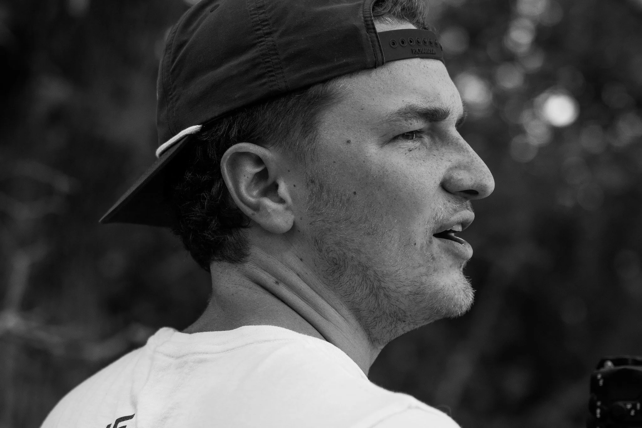 Bradley Gossett in profile view wearing a backward baseball cap, with a focused expression, set against a blurred outdoor background.