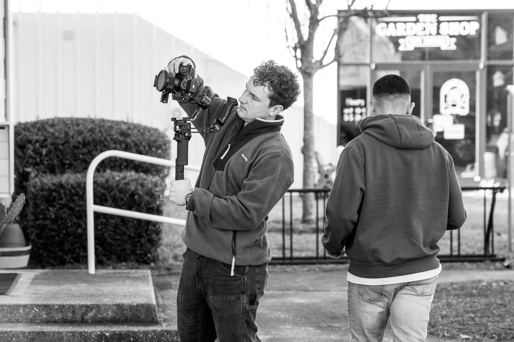 Bradley Gossett operating a camera on a stabilizer while Nick Rizzo stands beside him with their back turned, outside near a garden shop in Wilmington.