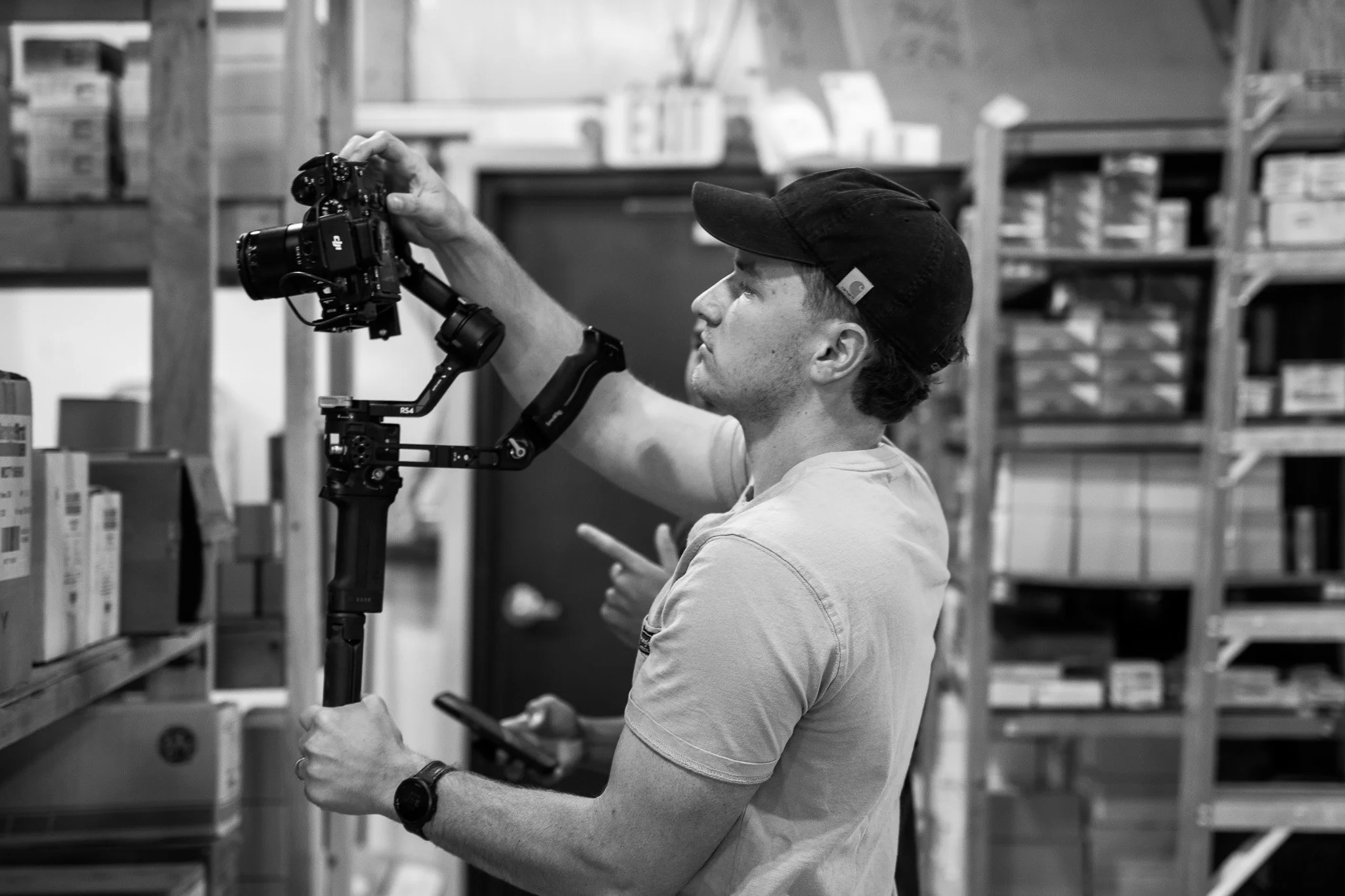 Bradley Gossett with a cap operating a camera mounted on a stabilizer or gimbal in a warehouse, holding a smartphone in his other hand.