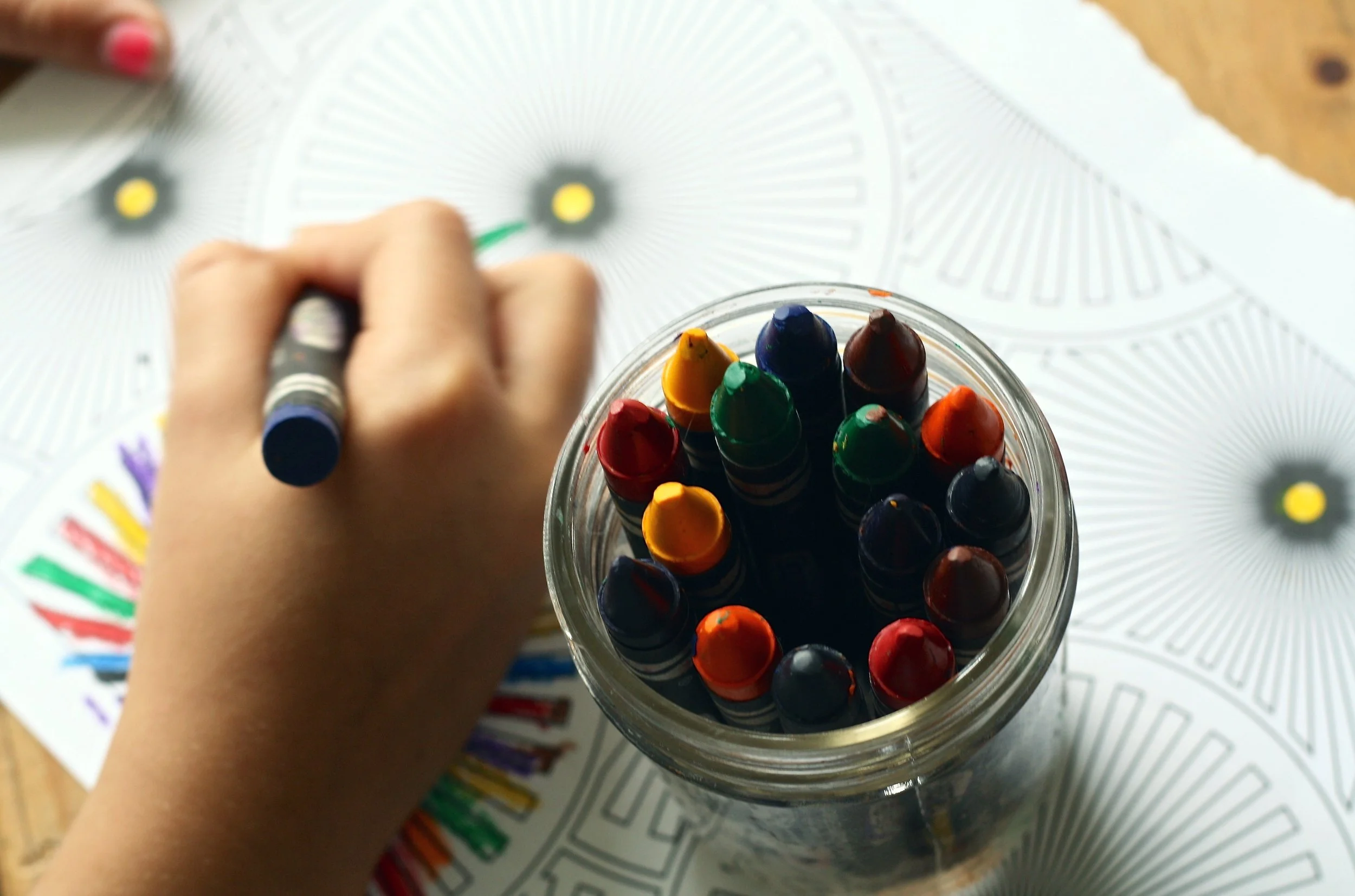 A hand holding a crayon over a container of gel pens on a white surface with a colorful trending example printed underneath.