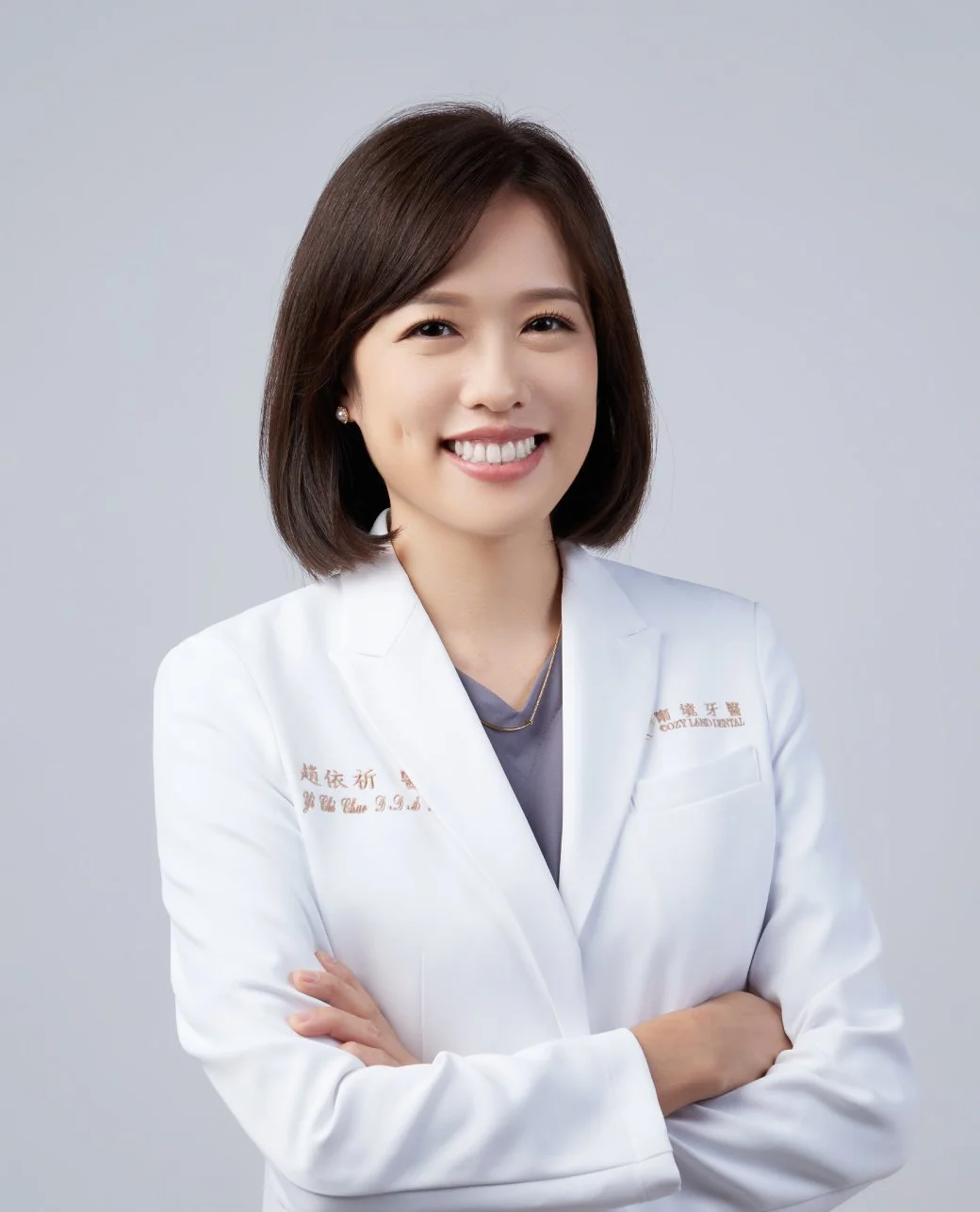 Smiling woman with shoulder-length dark hair wearing a white coat with Chinese characters and English text, crosses arms, against plain gray background.