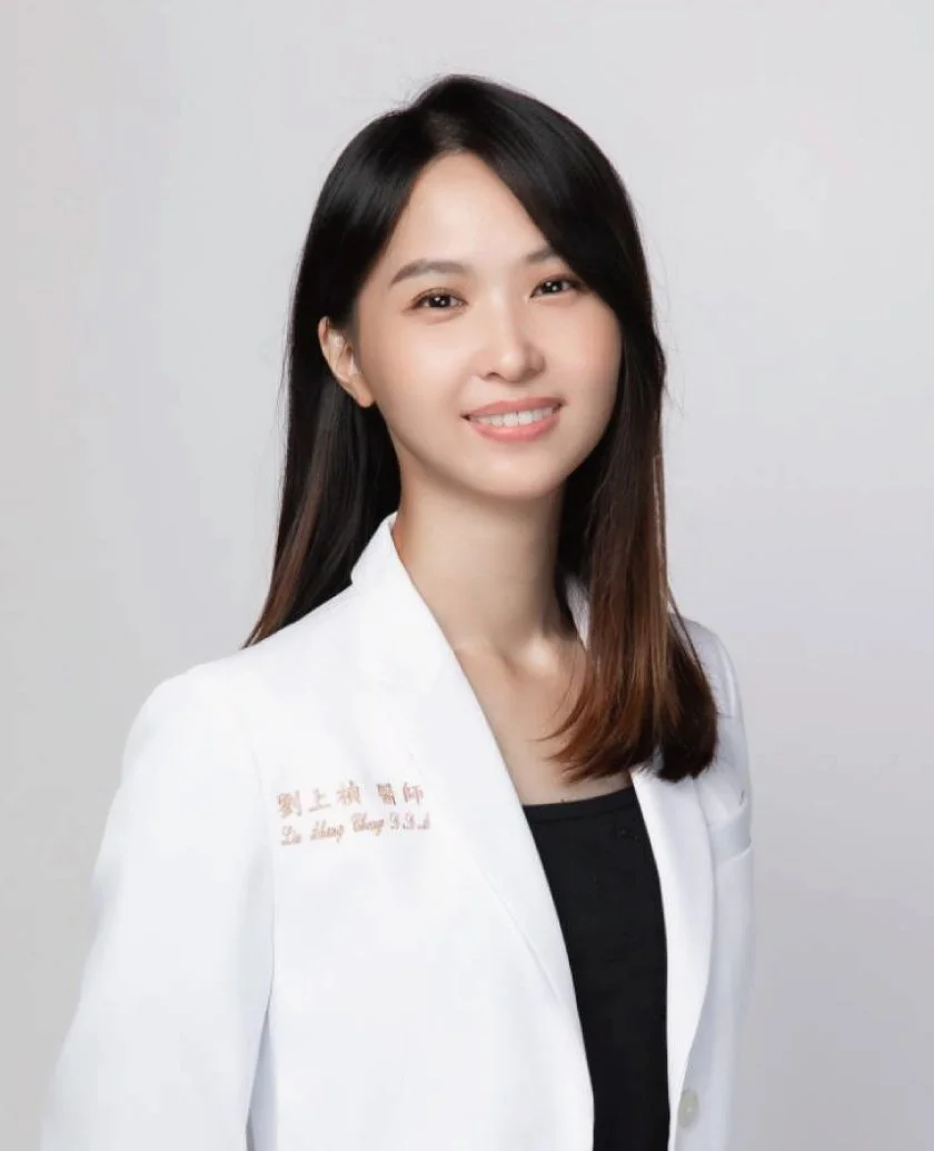 A young Asian woman with shoulder-length dark hair, wearing a white coat with red embroidery, standing against a plain light background, smiling gently at the camera.