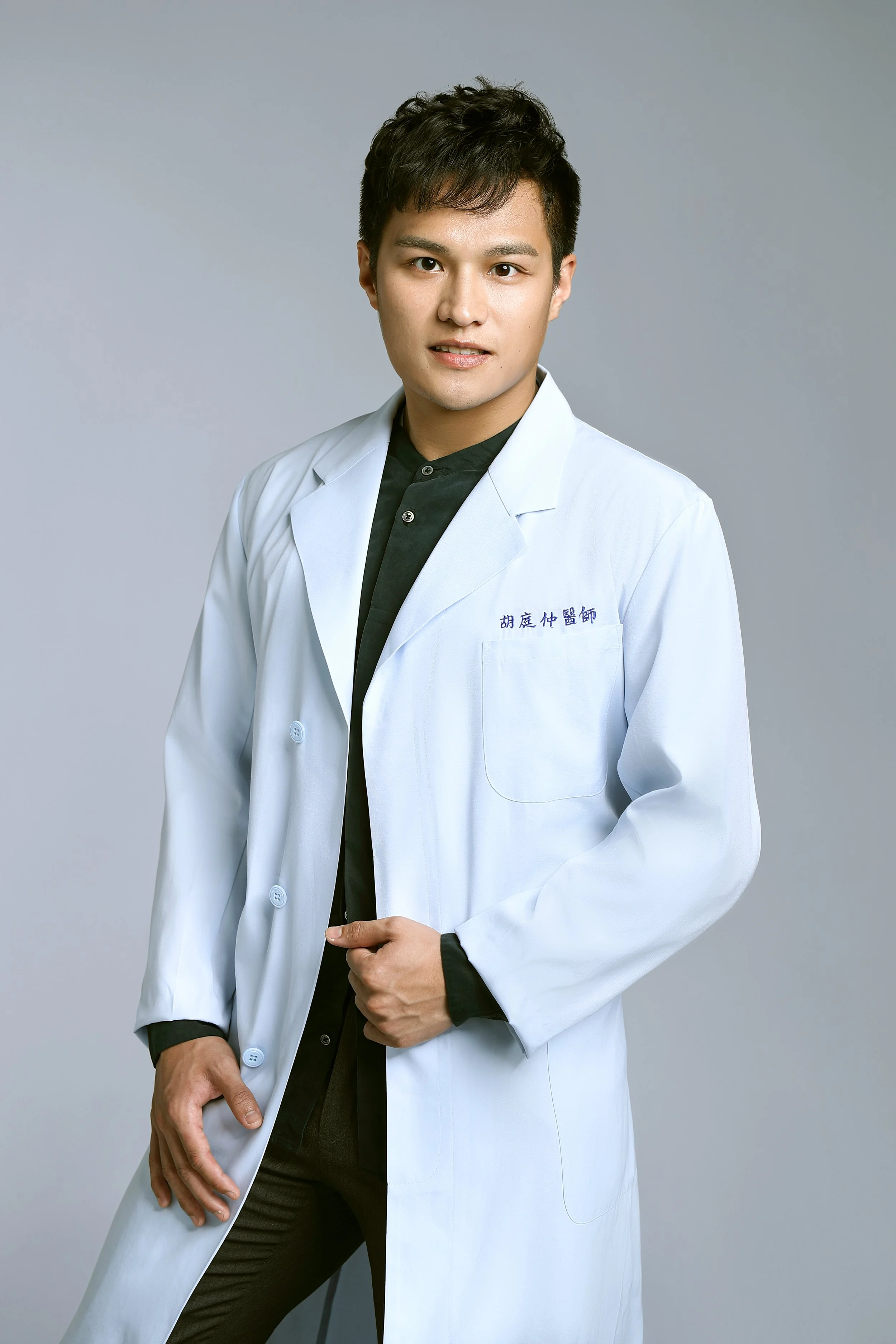 A young male doctor with short dark hair, wearing a white coat over a black shirt, standing against a plain light gray background.