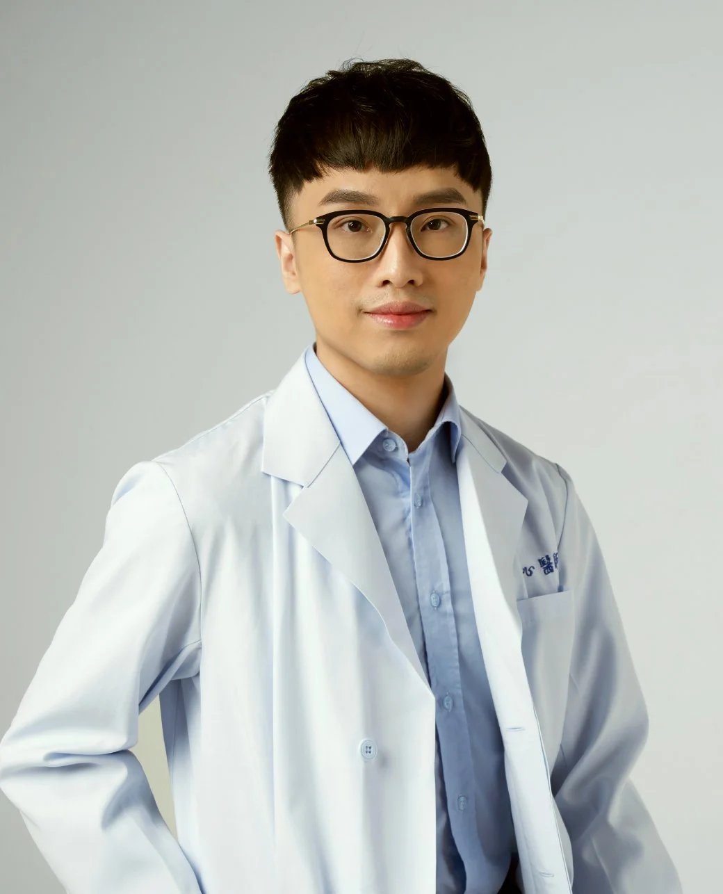 A young Asian male doctor with short black hair, wearing black-rimmed glasses, a light blue dress shirt, and a white medical coat, standing against a plain light gray background.