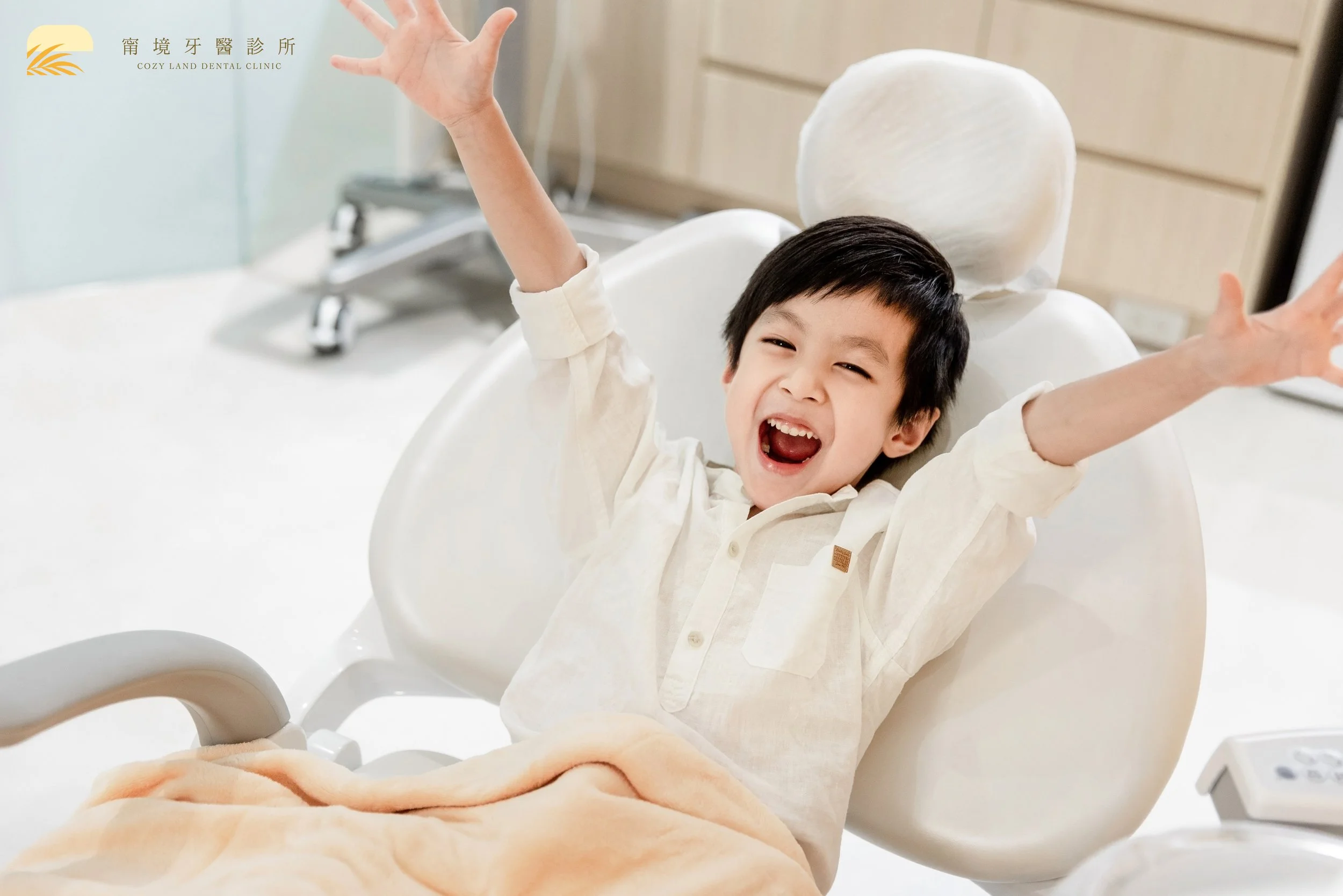 A young boy sitting in a dental chair with arms raised, smiling widely in a dental clinic.