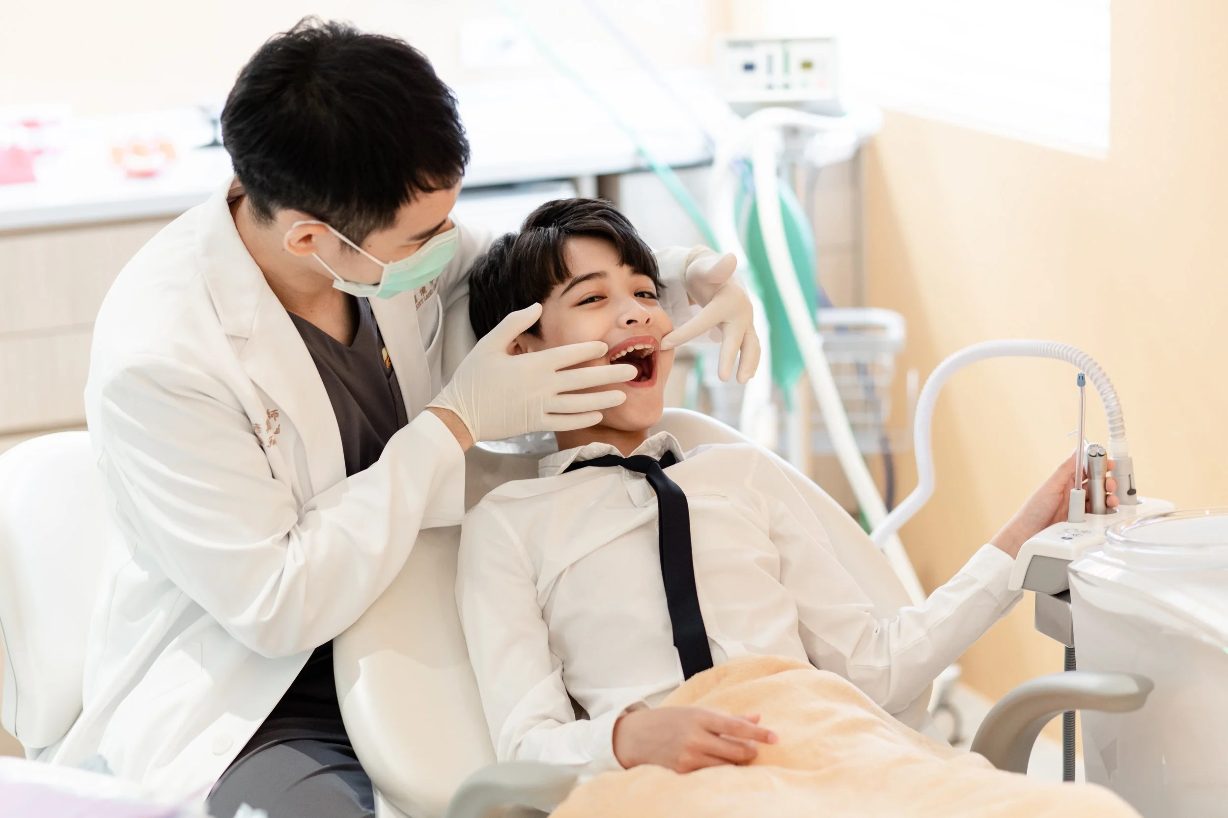Dentist examining a young boy's teeth in a dental office with dental equipment in the background.