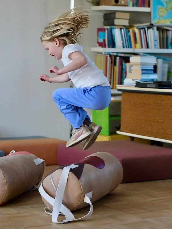 A child jumps over a punching bag lying on the floor and laughs.