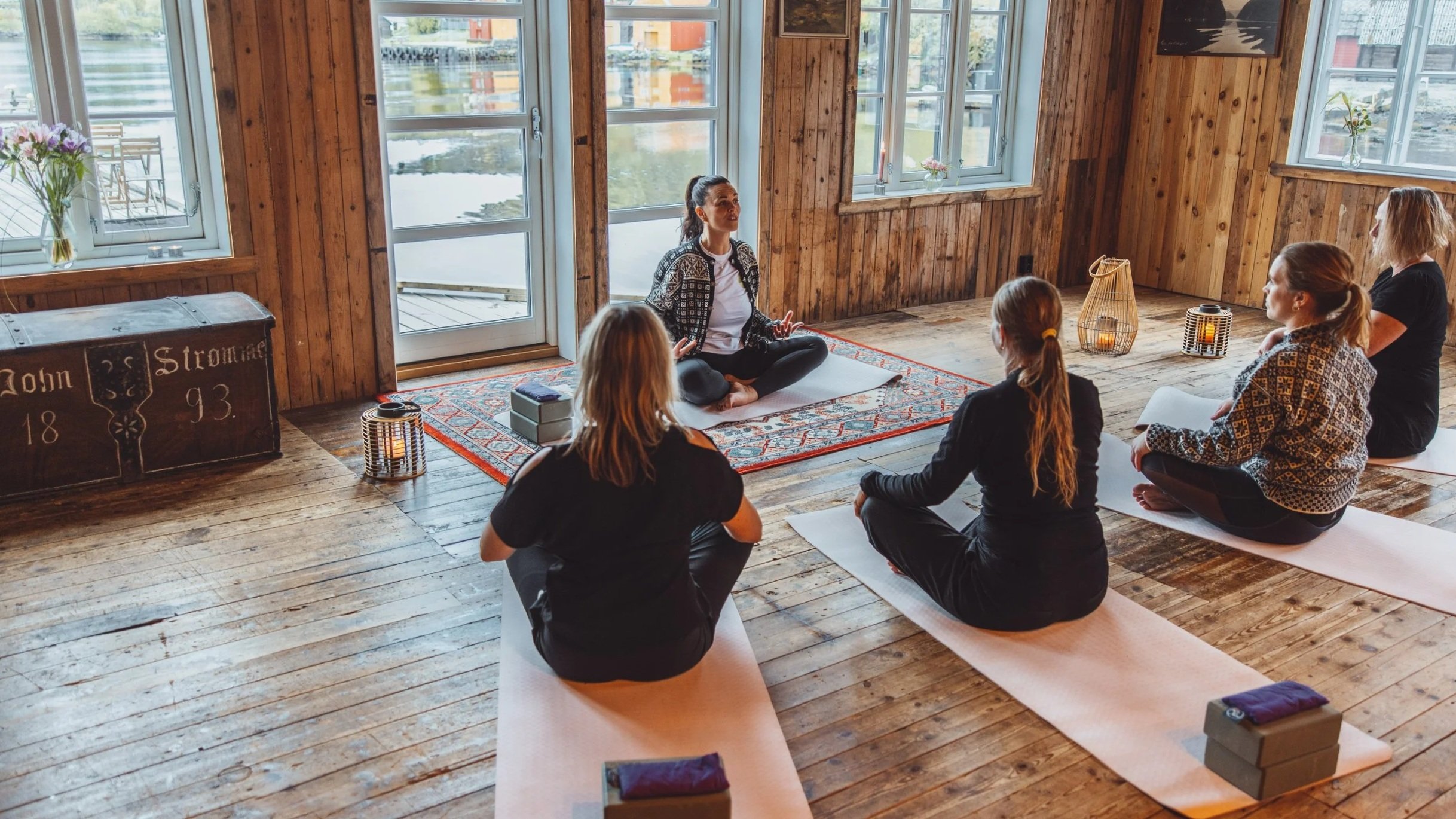 En gruppe mennesker deltar i en yoga-klasse i et rom med trevegger og store vinduer, hvor de sitter på yogamattaer i en sirkel rundt en instruktør som sitter på en matte.