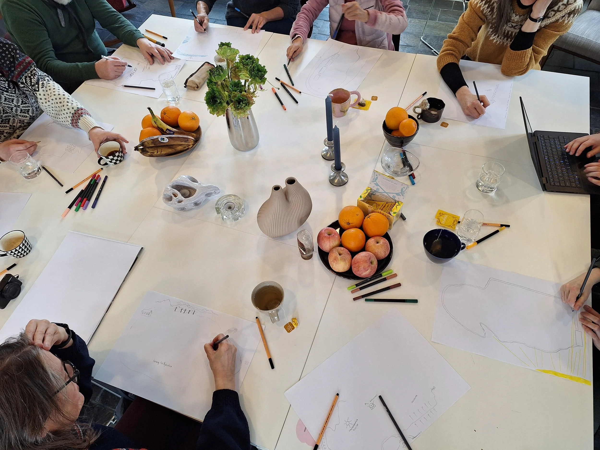 En gruppe mennesker som sitter rundt et bord og holder på med aktiviteter som farging og tegning. På bordet er det også en vase med blomstrer, lysestaker og noen fruktskåler.