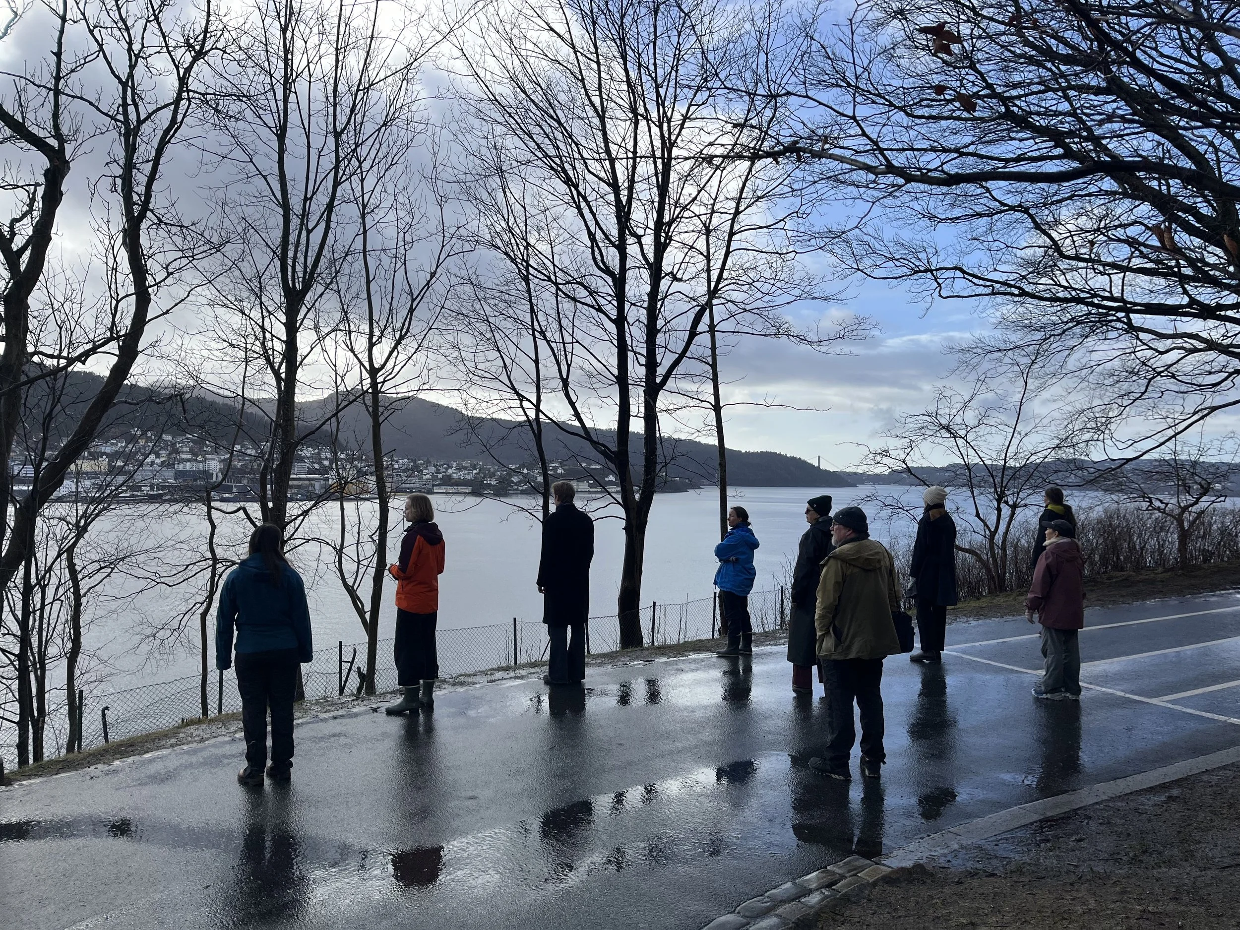 En gruppe mennesker står på en asfaltvei og lytter og ser ut mot et vann
