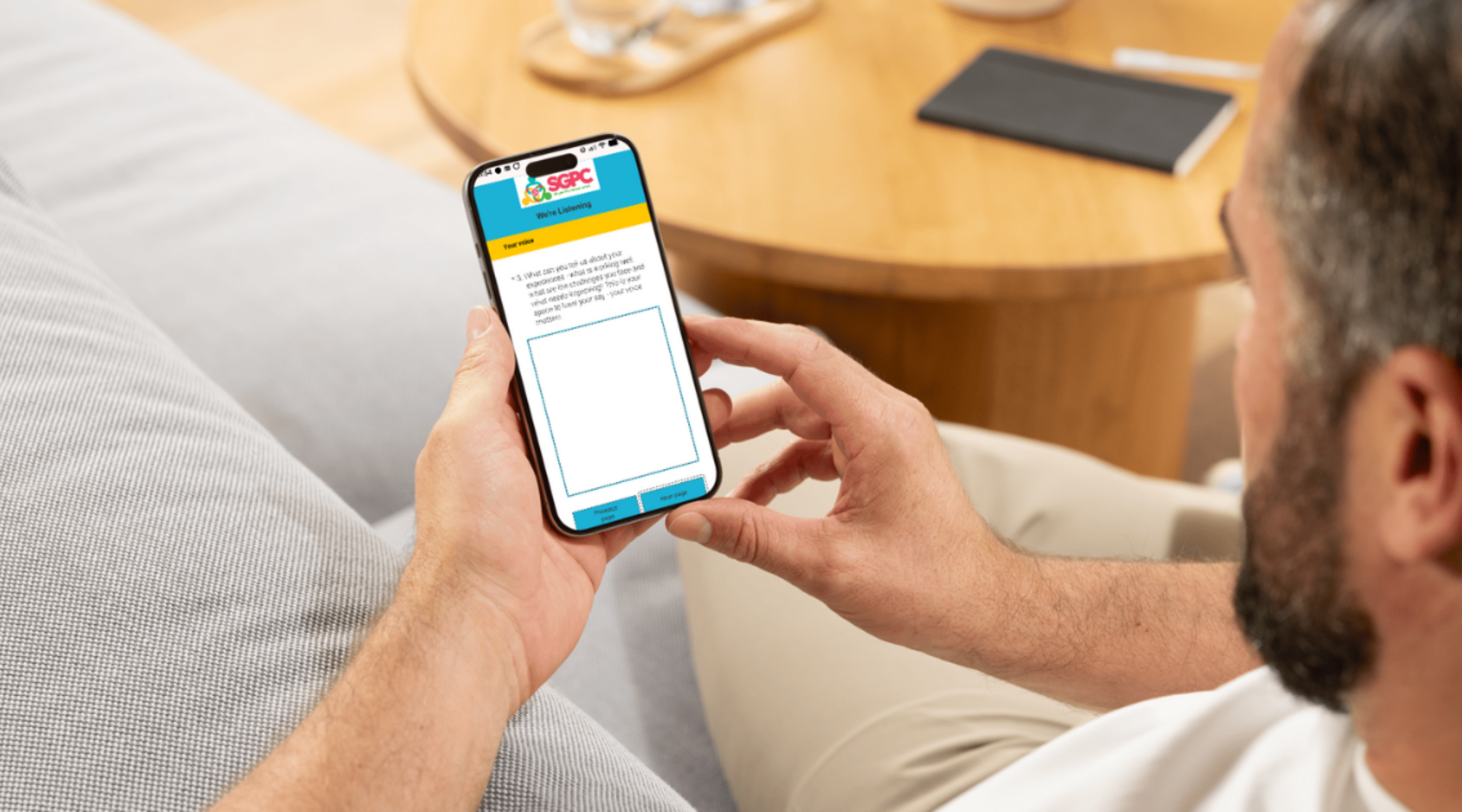 Man sitting on a sofa holding his phone, the screen with the SGPC logo and a text entry box is visible.