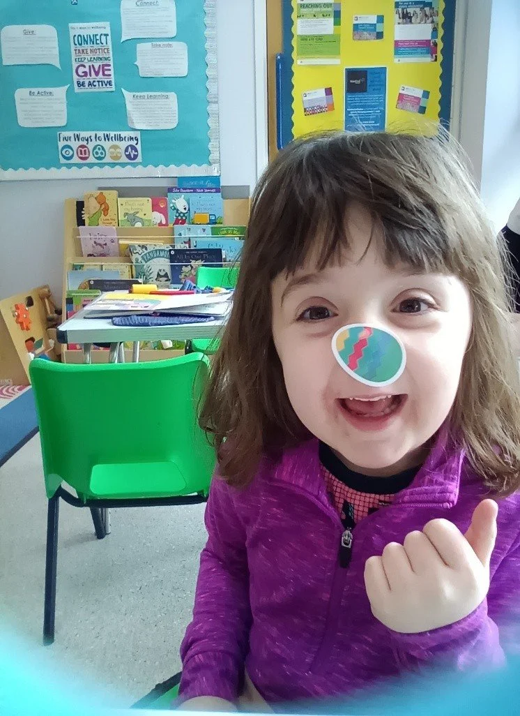 Young girl wearing a pink top is smiling into the camera. A colourful easter egg shaped sticker is stuck on her nose.