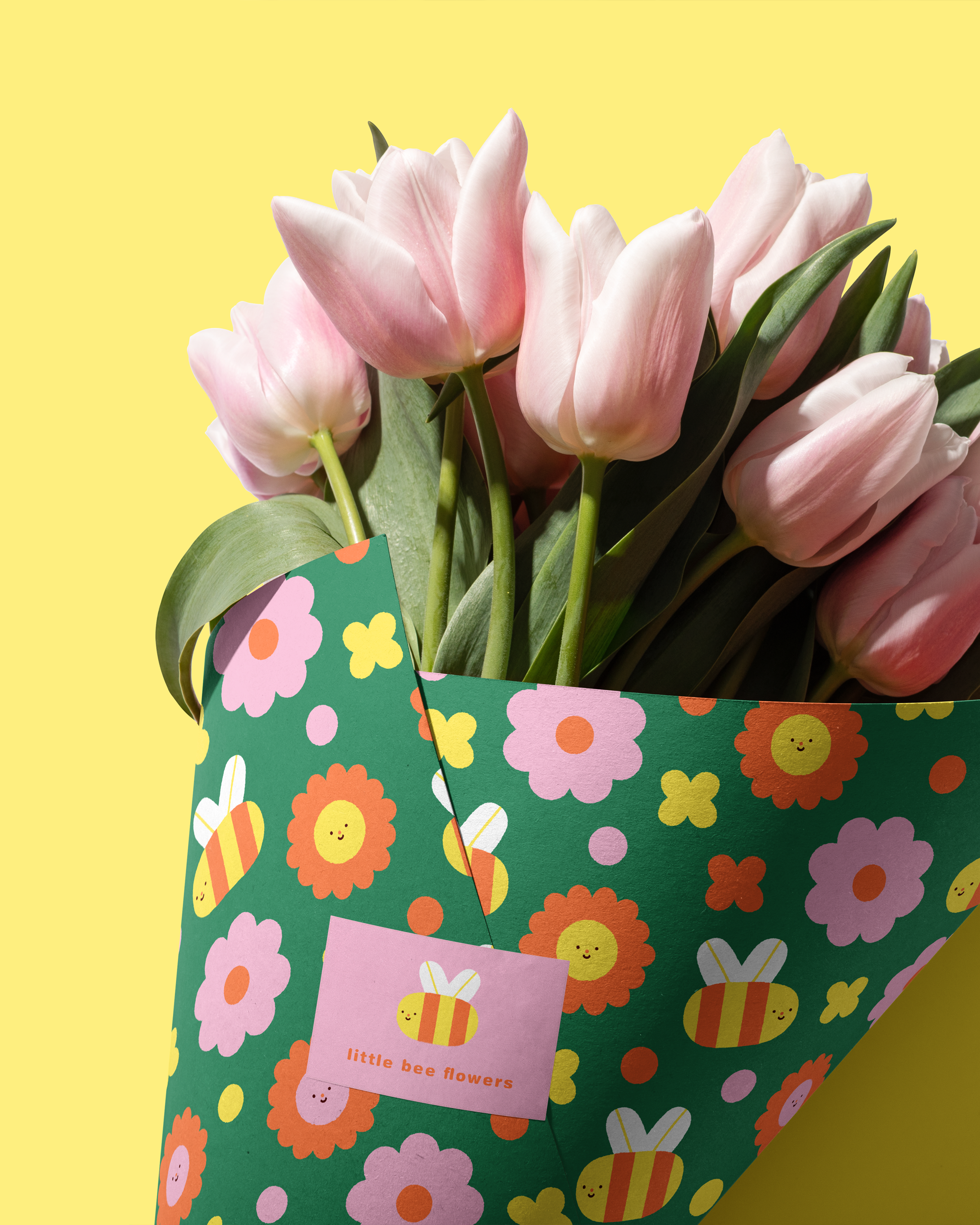 A bouquet of pale pink tulips in a decorative gift bag with colorful flower and bee patterns, and a pink tag reading 'little bee flowers'.