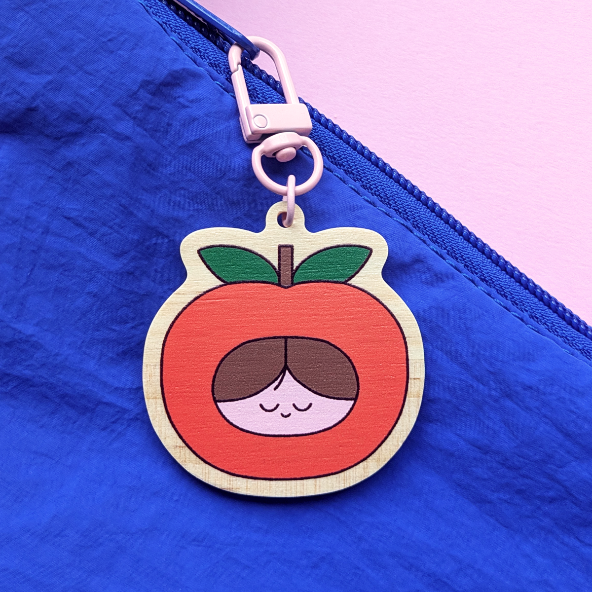 A wooden keychain hanging from a blue zipper on a fabric surface, with a cartoon face of a girl inside an orange fruit with green leaves.