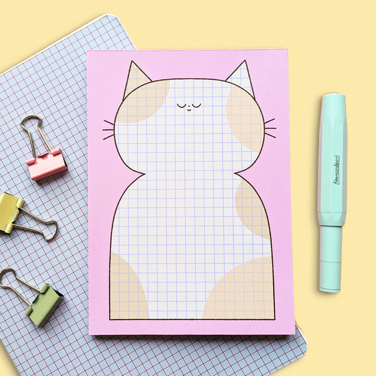 Decorative paper featuring a cute, minimalist cat illustration with a pink background, surrounded by stationery items including pastel-colored binder clips and a mint green marker, set on a pastel-colored surface.