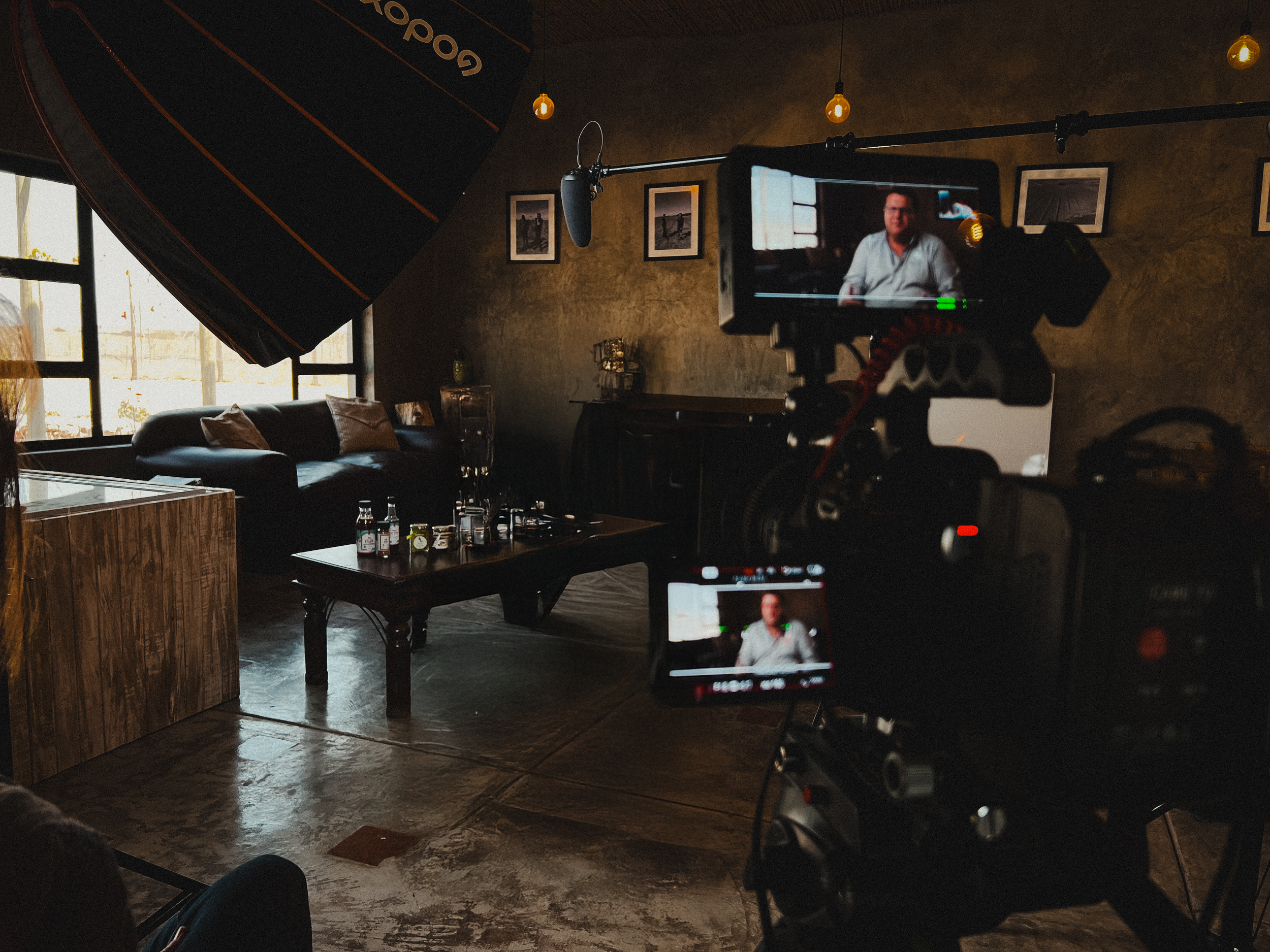 A person is being filmed on a professional camera in a dimly lit room with large windows, a dark wall decorated with framed photos, a black sofa with pillows, and a coffee table with bottles and glasses.