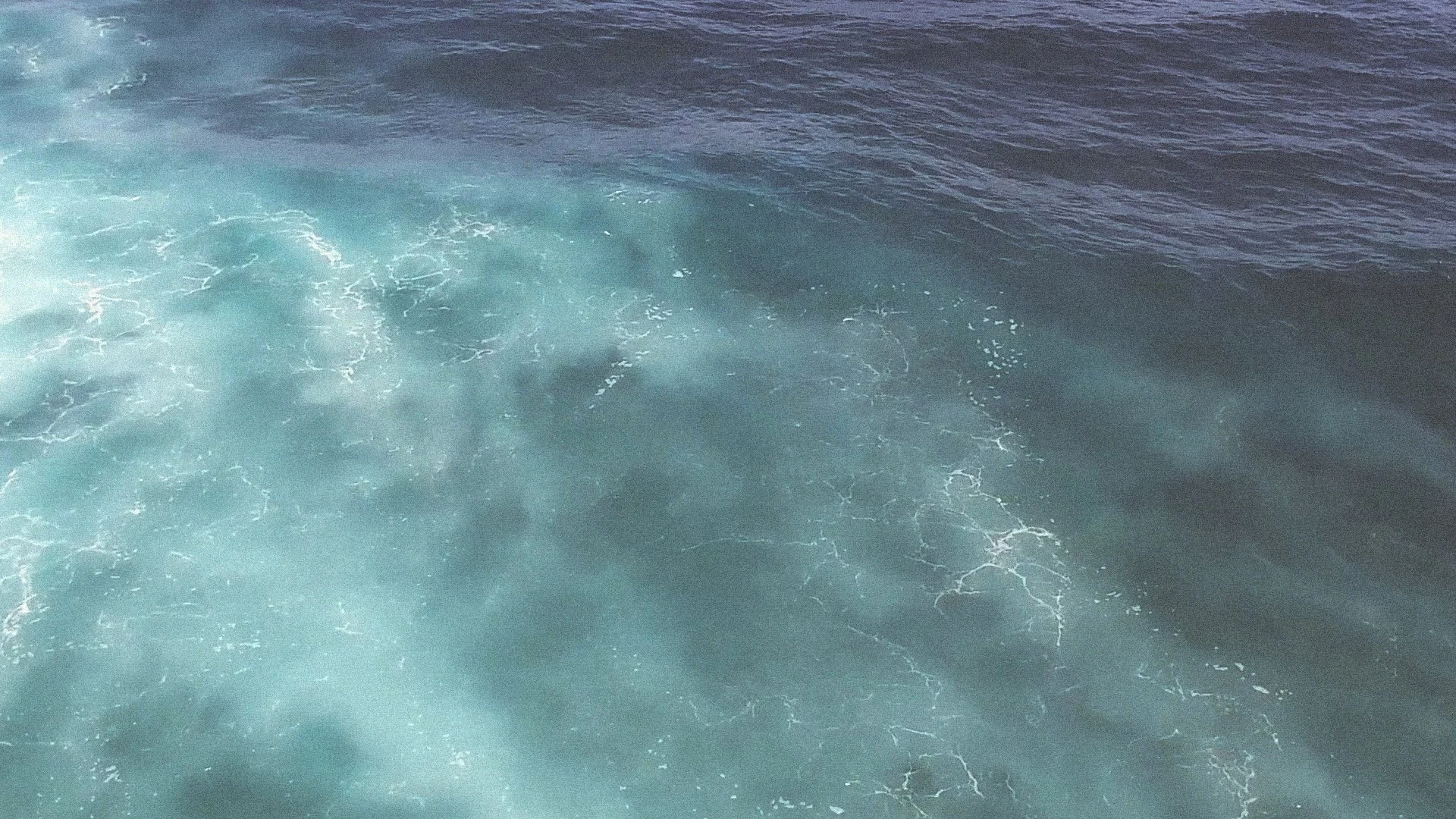 De foto toont de oceaan met verschillende tinten blauw en lichtblauw, met golven en wit schuim op het wateroppervlak.
