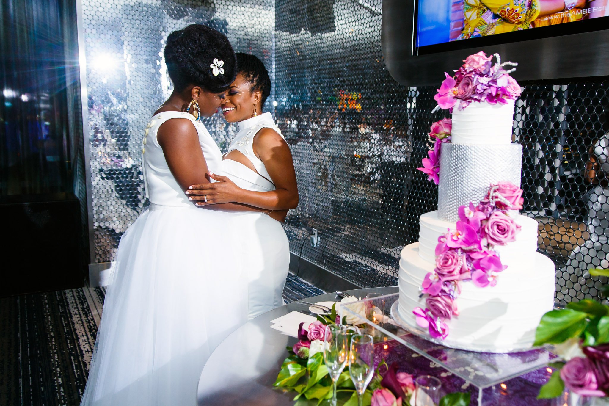 Queer brides in white wedding attire lean in to kiss by their cake in Dallas Texas The Amber Studio
