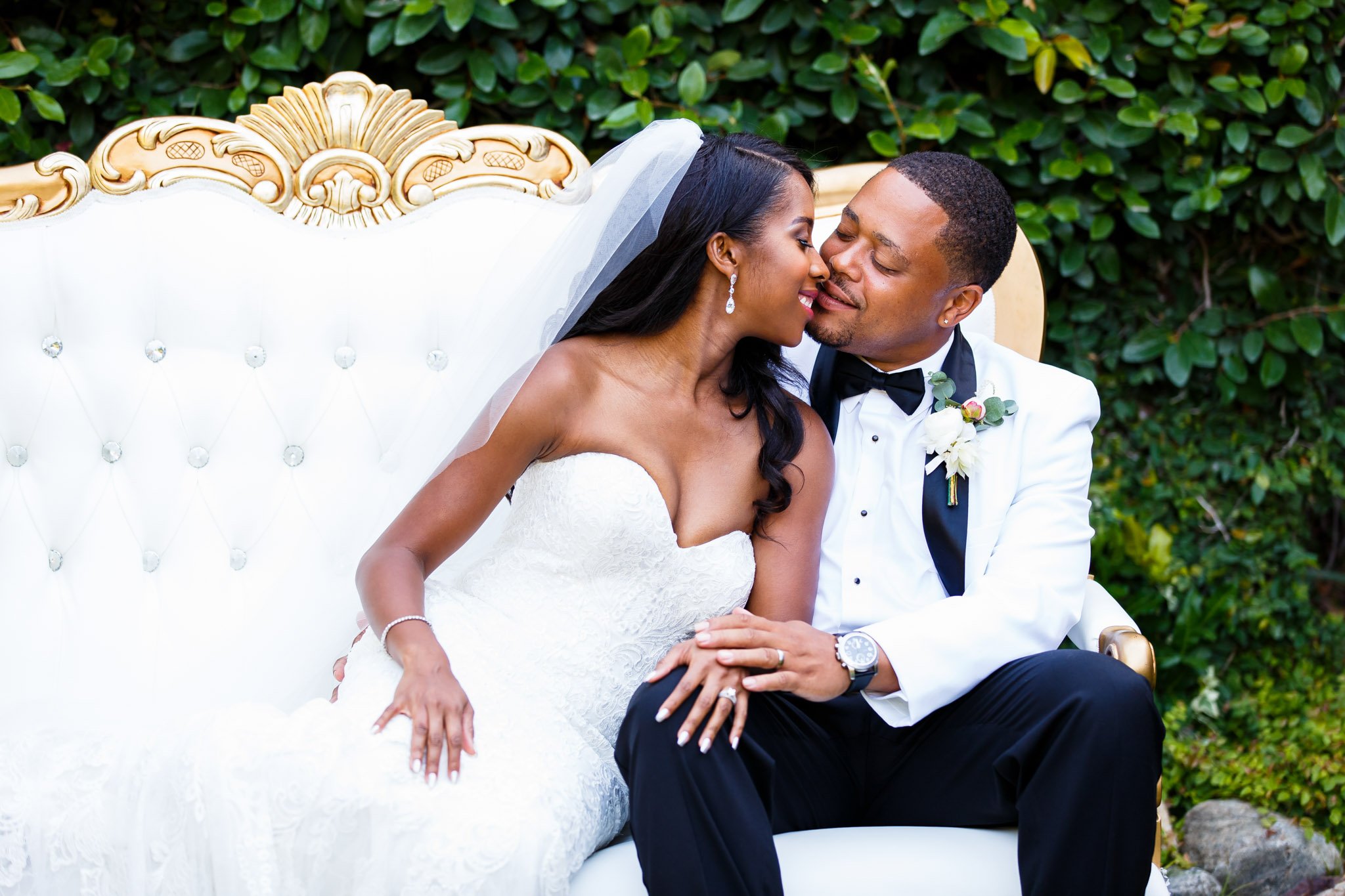 A newlywed couple in wedding attire lean in to kiss on a white couch outside in Texas The Amber Studio