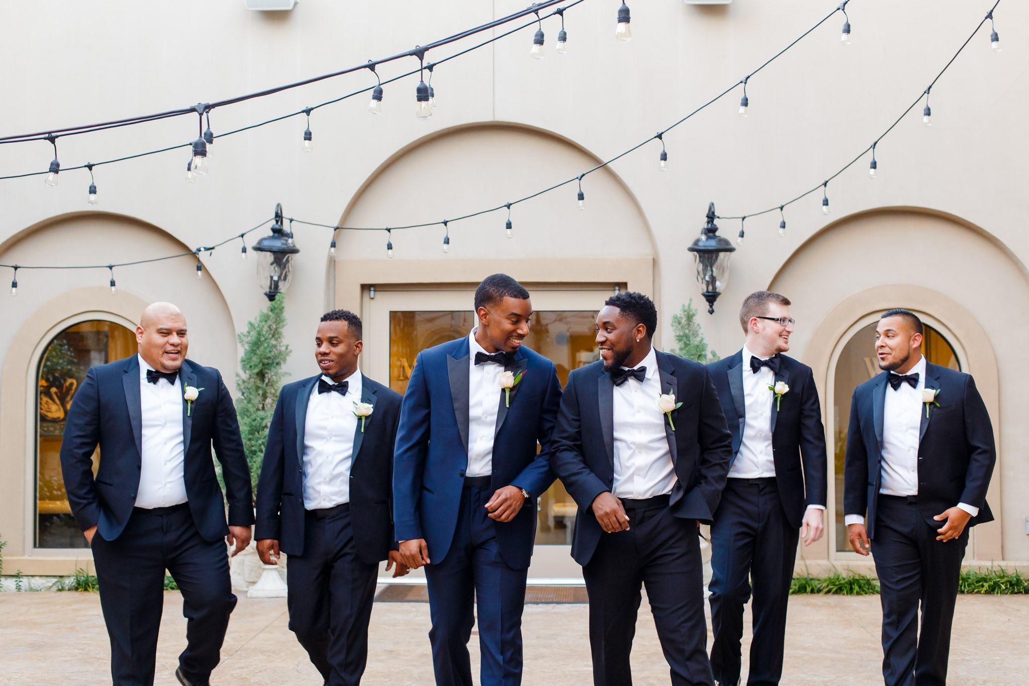 A groom and his groomsman walking and smiling under string lights in Dallas Texas The Amber Studio