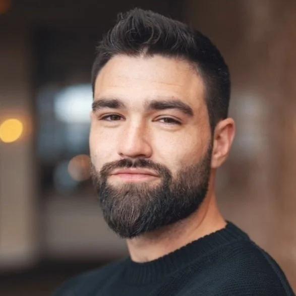 A man with dark hair, a full beard, and wearing a black sweater, standing indoors with a blurred background featuring warm lighting.