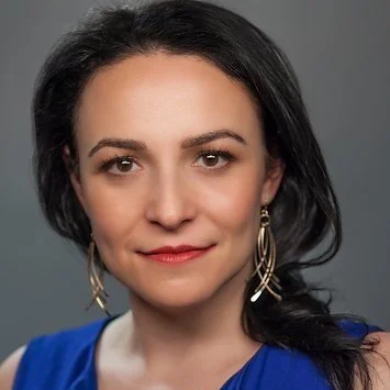 A woman with dark hair wearing a blue top and gold hoop earrings, looking directly at the camera with a neutral expression.