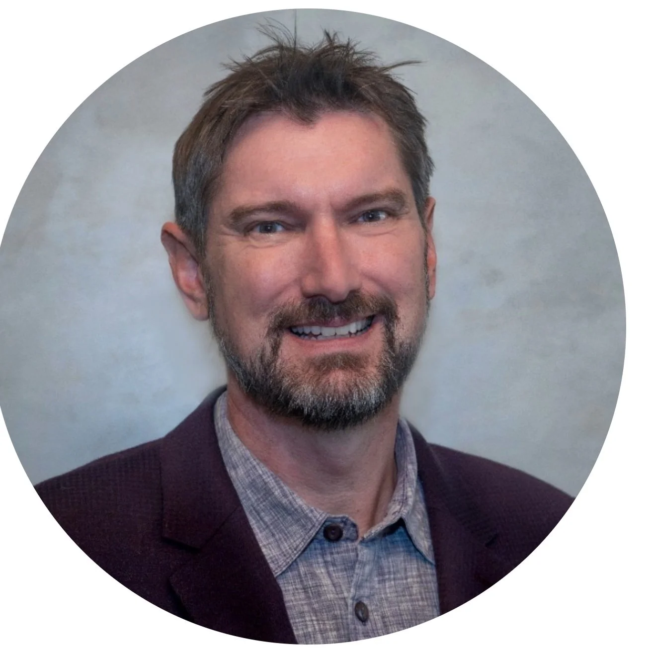 A smiling man with brown hair and a beard wearing a checked shirt and a dark blazer, against a neutral background.