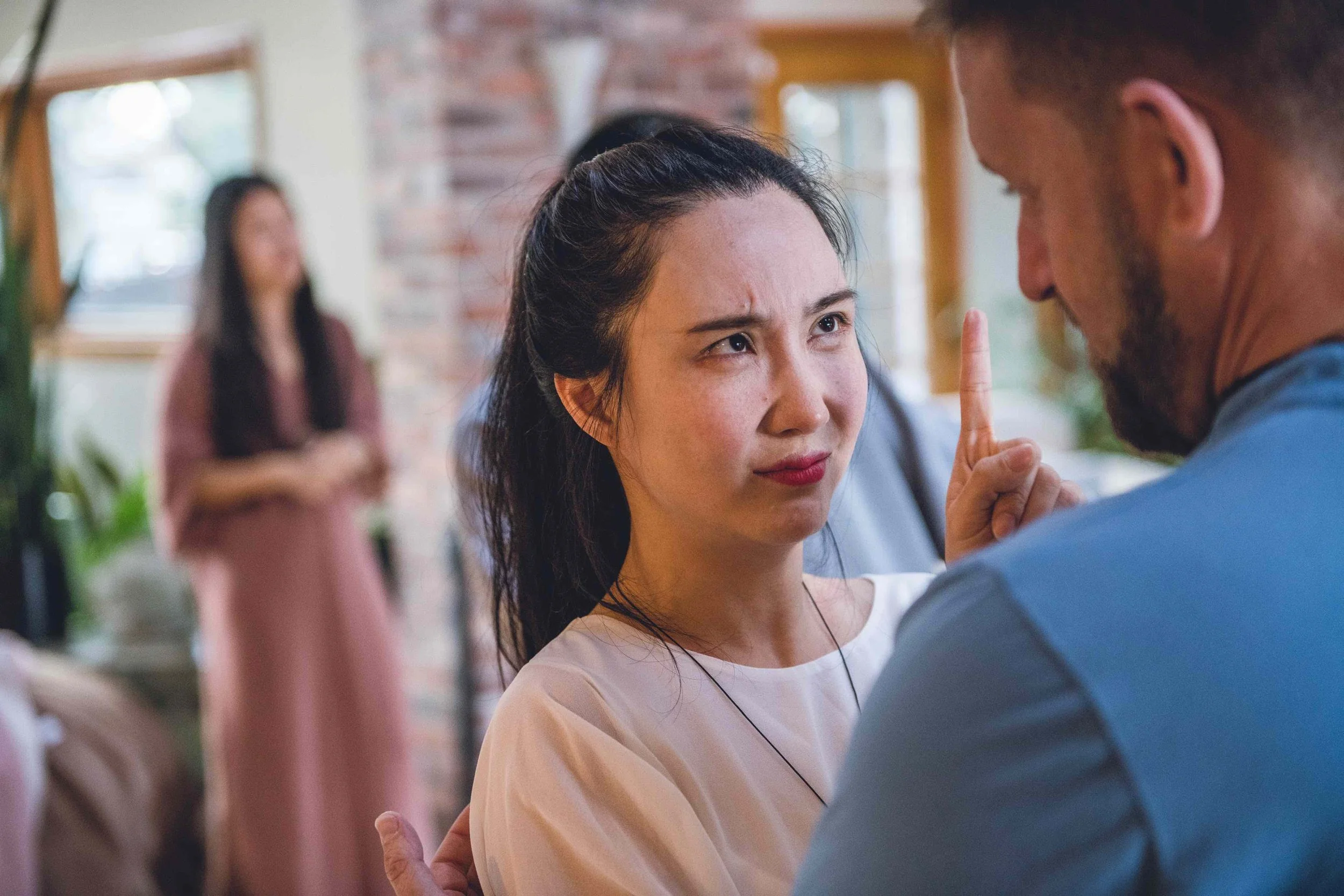 A woman with dark hair pulled back, making a stern face, has her index finger raised and is facing a man with a beard and short hair in a blue shirt. In the background, a woman in a pink dress is blurred near a window with brick walls and plants.