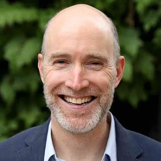 A smiling middle-aged man with a beard and bald head, wearing a suit jacket and shirt, standing outdoors with greenery in the background.
