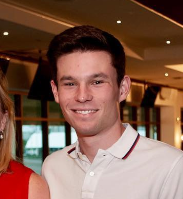 A young man with short dark hair, wearing a white polo shirt with a striped collar, smiling in an indoor setting with warm lighting.