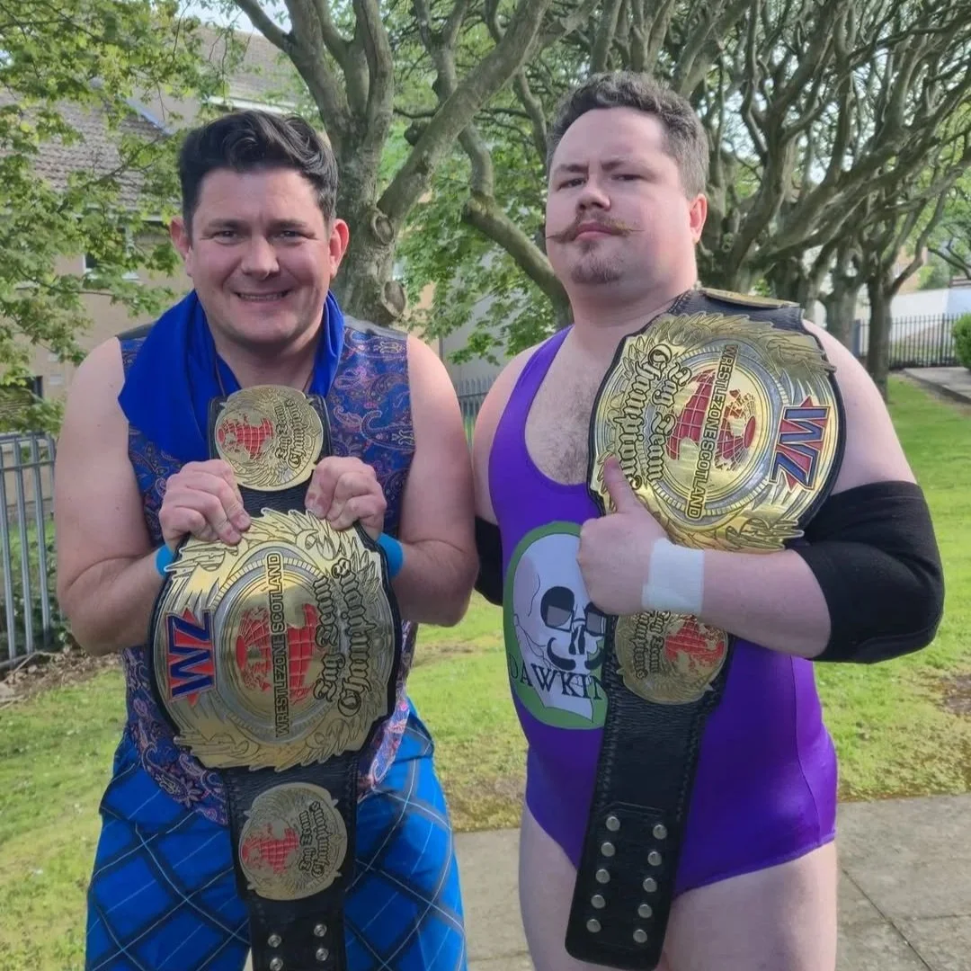 Two men posing with championship wrestling belts. The man on the left has a mustache, curly hair, and wears a purple wrestling singlet. The man on the right has dark hair, a purple scarf, and a sleeveless vest. Both are smiling at the camera.