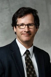 A man with dark hair and glasses wearing a suit and tie, smiling against a gray background. David Forliti, Dave Forliti, UST