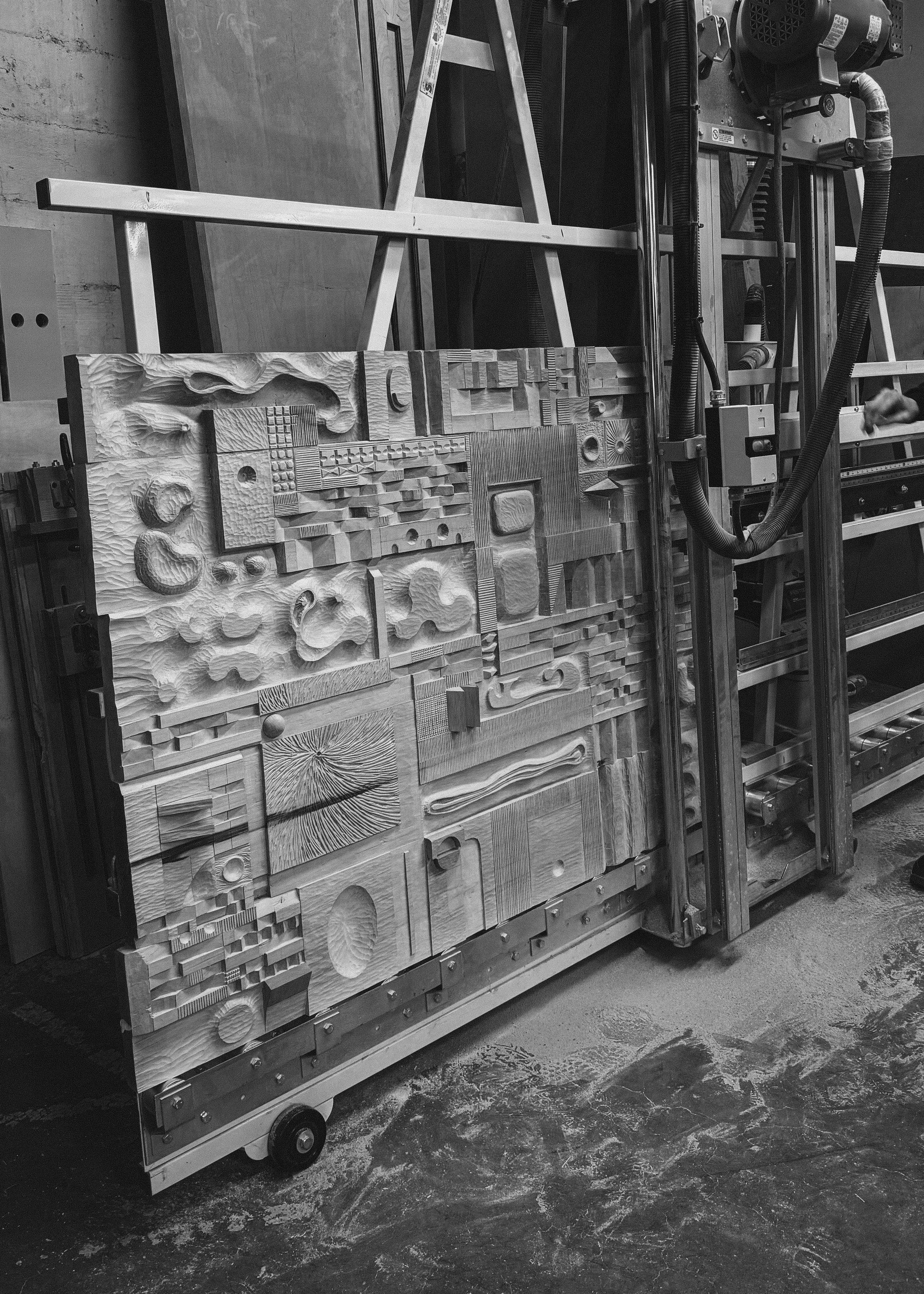 A large-scale, hand-carved wood relief panel by Agata Rek, featuring a complex grid of geometric and organic textures, mounted on a vertical studio rack.