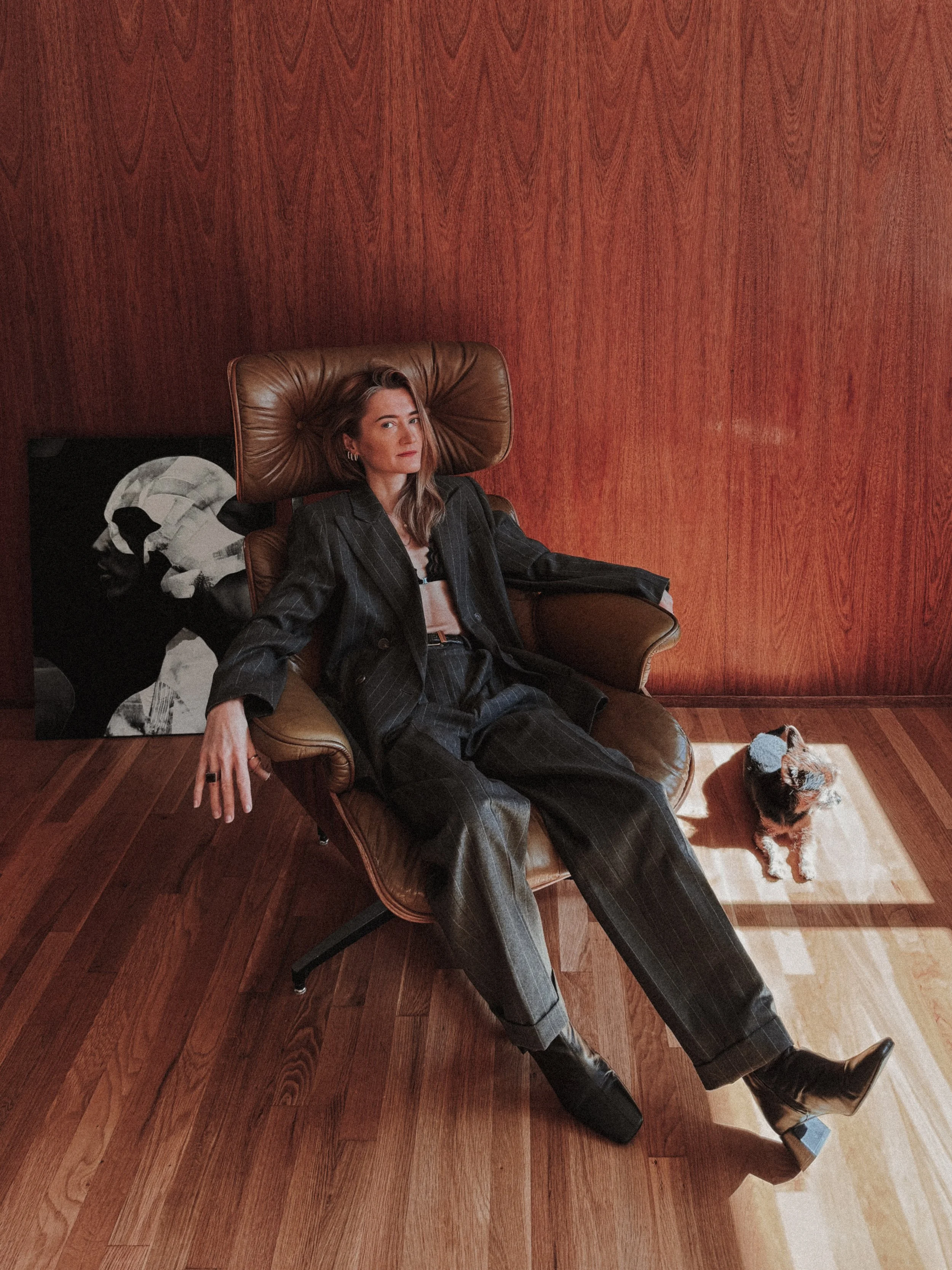 Contemporary wood sculptor Agata Rek sitting in a leather Eames-style lounge chair in a wood-paneled studio, wearing a charcoal pinstripe suit, with a small dog and abstract artwork in the background.