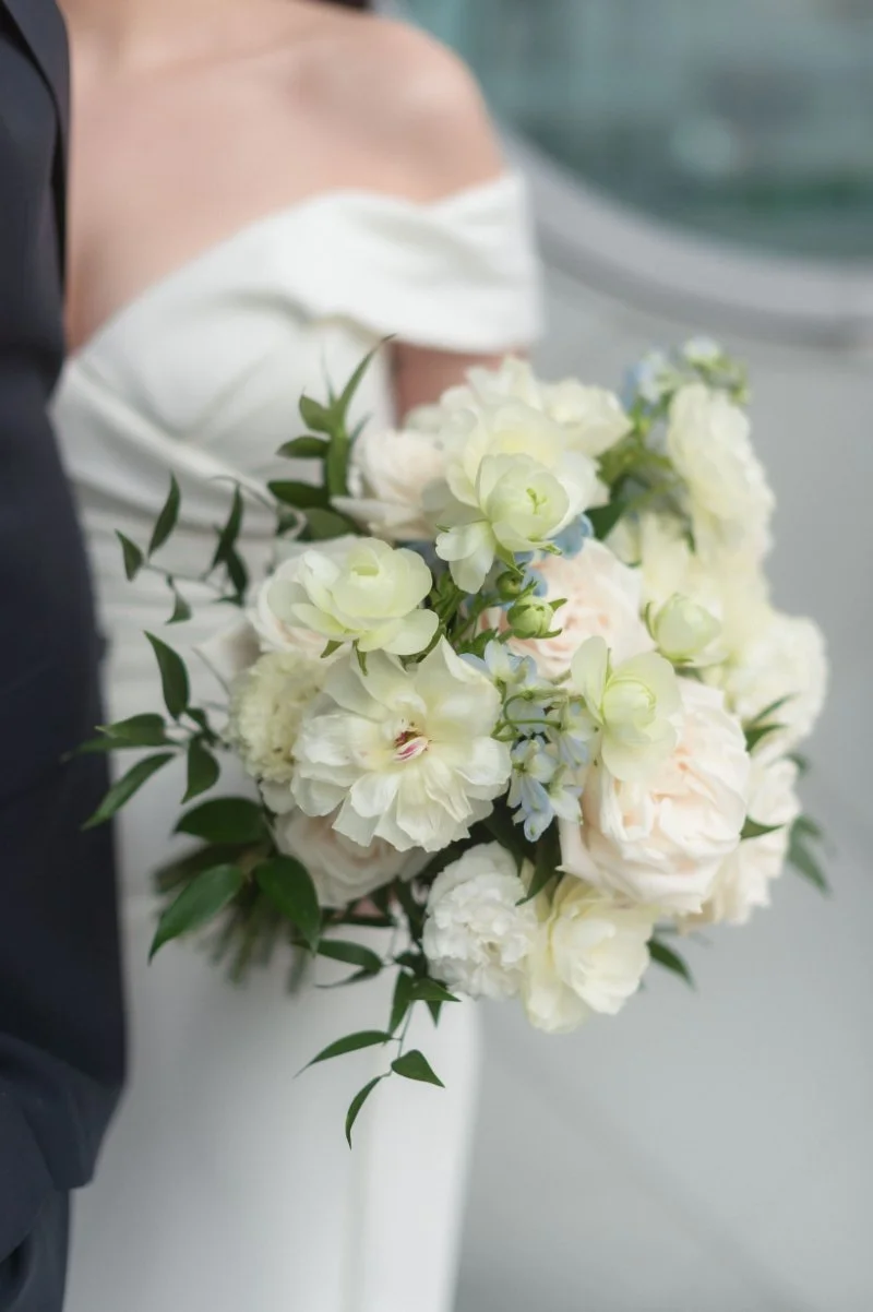 Soft ivory and blush bridal bouquet with greenery held against classic white gown.