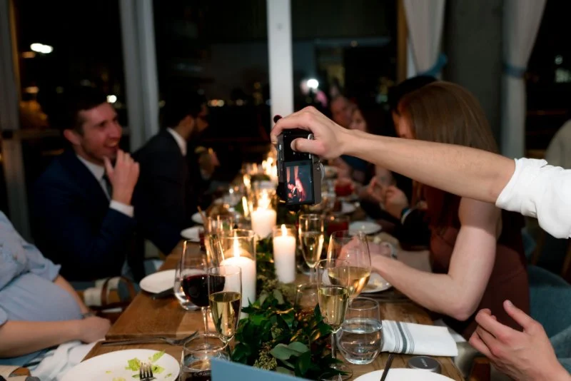 Friends gathered around a candlelit dinner table, laughing and taking photos during a warm evening celebration