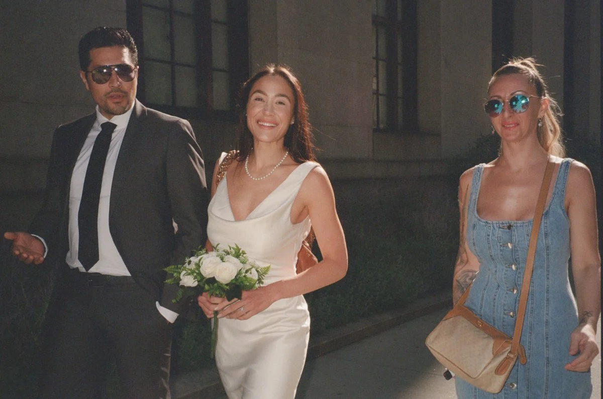 Bride in a white dress holds a small bouquet while walking outdoors between two companions near a stone building, captured in bright sunlight with a candid, timeless look typical of film wedding photography.