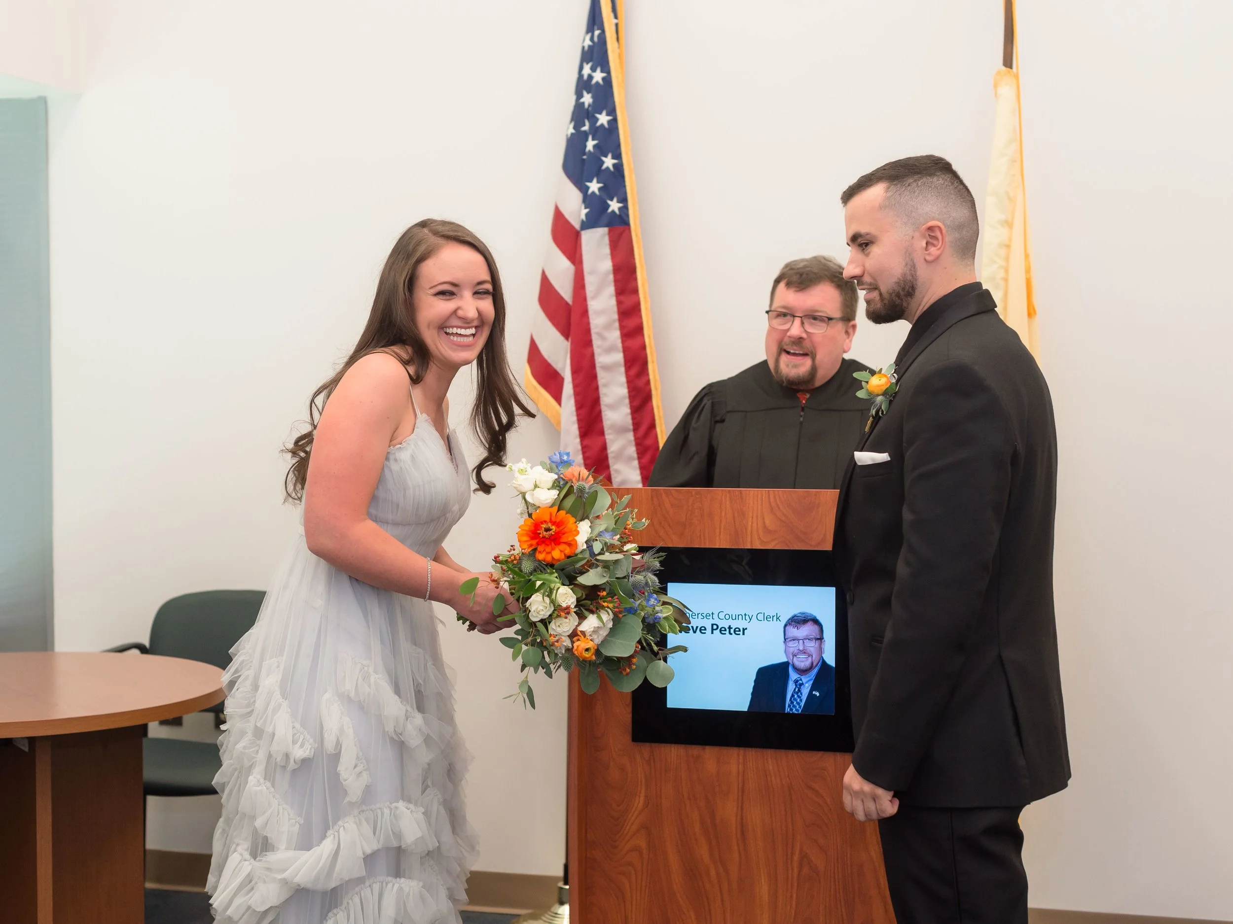  Couple smiles during their New Jersey City Hall wedding ceremony, standing before the officiant and exchanging vows in a heartfelt, intimate moment. 