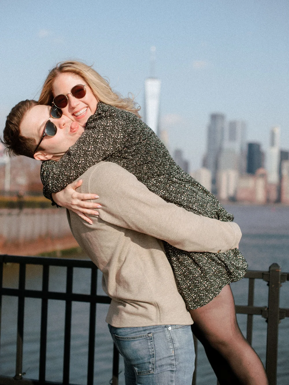  Couple embraces and laughs by NYC waterfront with skyline in the background.   