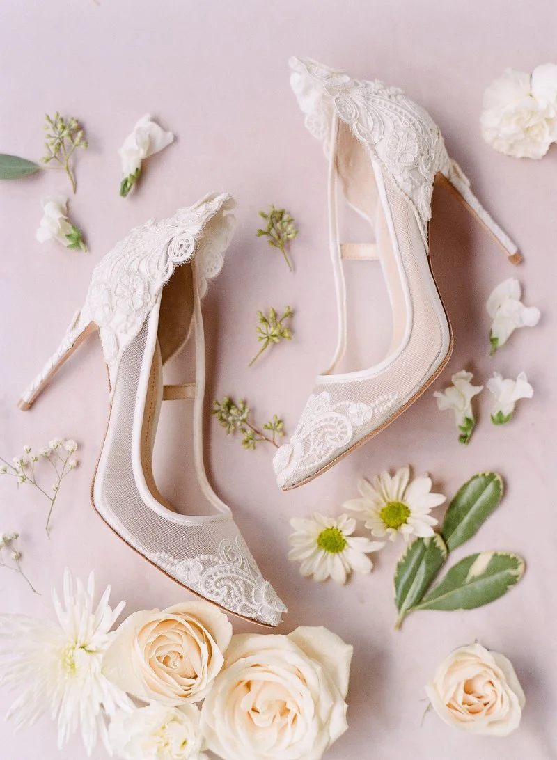  white flowers, greenery and bridal shoes flat lay against a pink background 