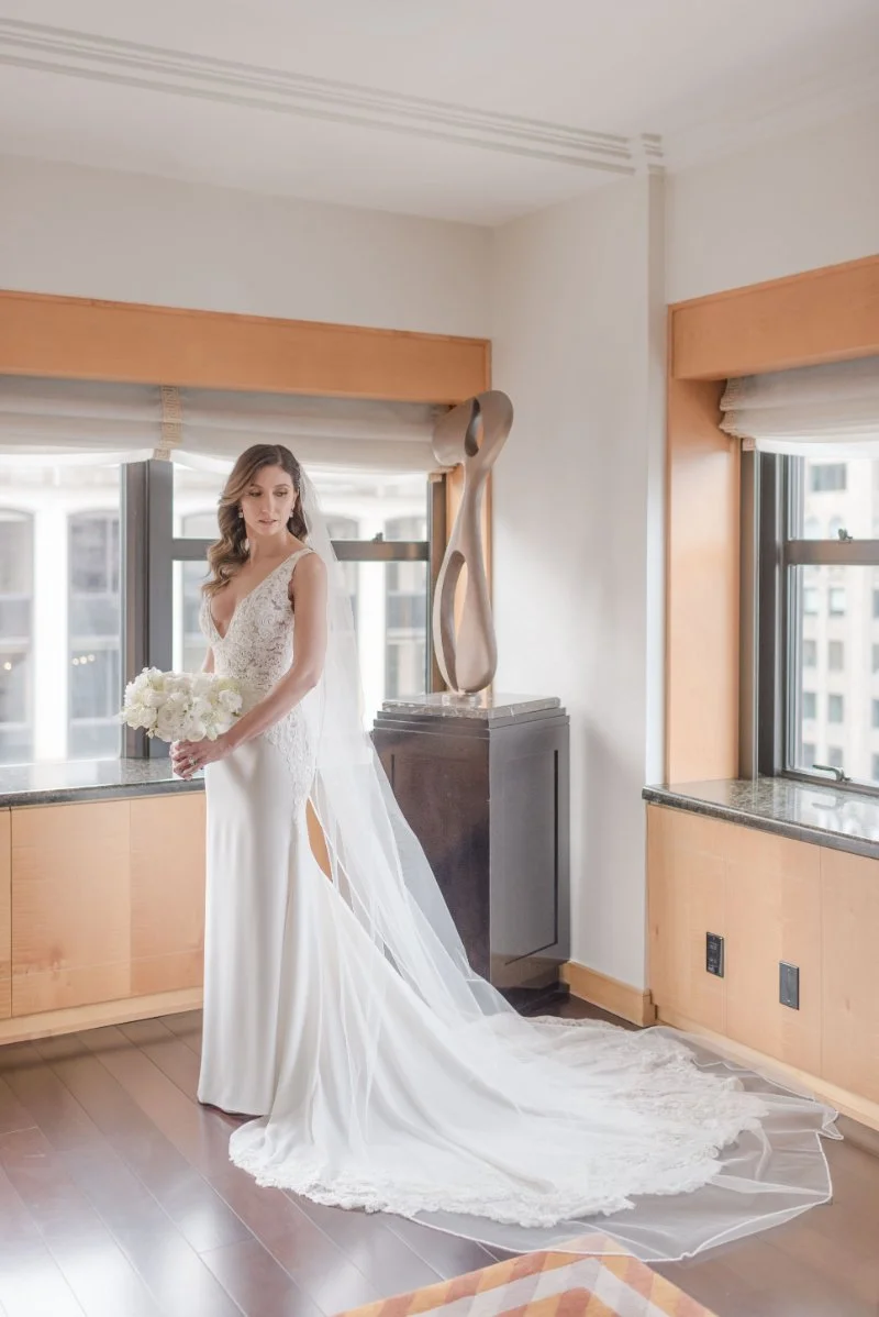  a bride standing in a bright lit window wearing her gown and veil holding her all white bouquet 