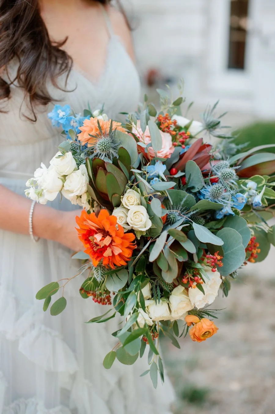  Close-up of a bride holding a colorful bouquet with orange, blue, and white blooms mixed with greenery — creative floral inspiration for winter wedding ideas that include vibrant seasonal flowers.   