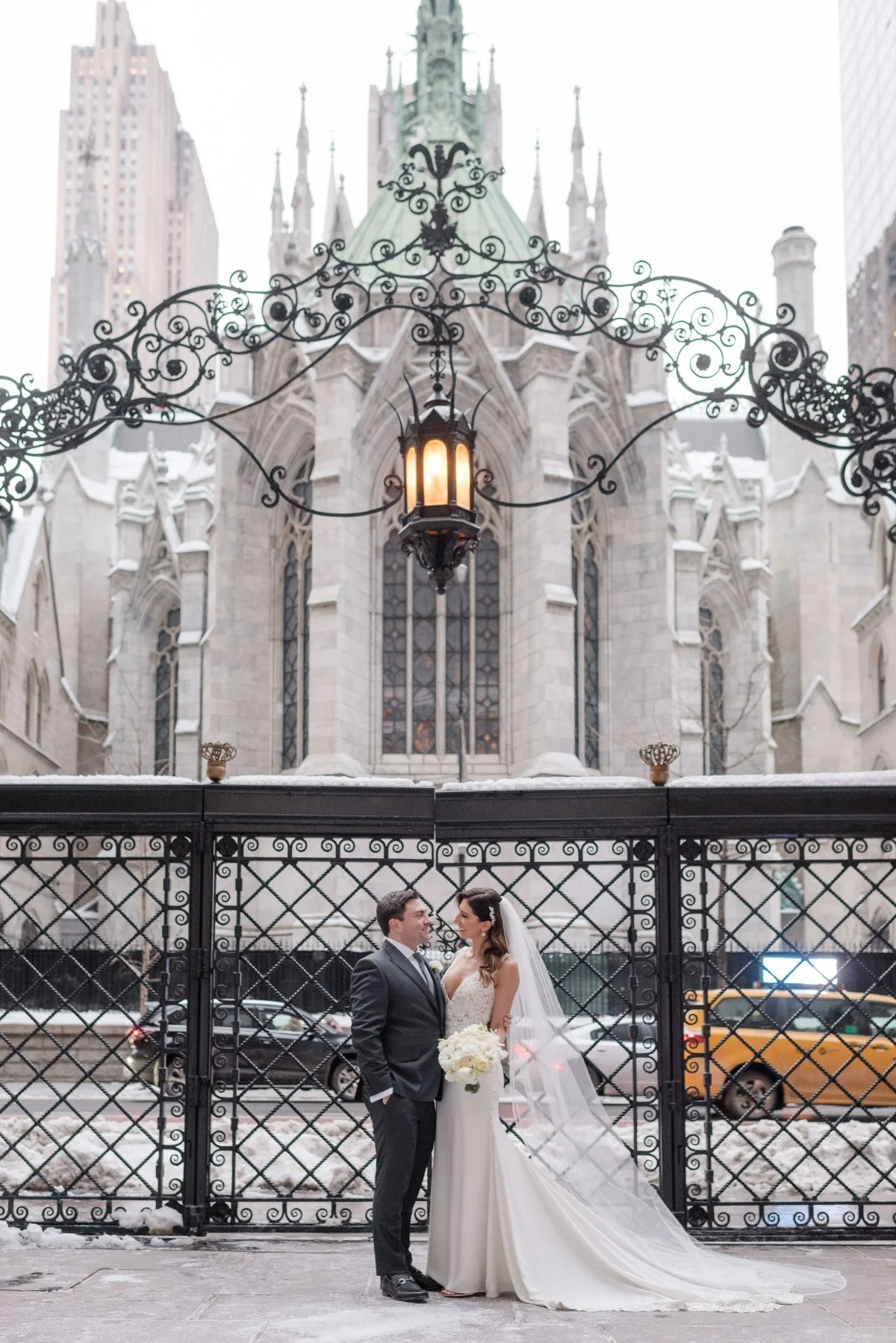  Bride and groom standing outside a grand stone church in the city on a snowy day, holding hands under an ornate iron gate with warm lantern light — a romantic example of winter wedding ideas in Manhattan. 