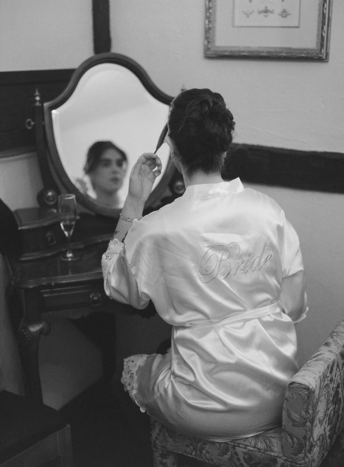   Black and white photo of a bride in a satin robe with “Bride” embroidered on the back, sitting at a vintage vanity mirror while styling her hair before the ceremony. A champagne glass sits nearby.    