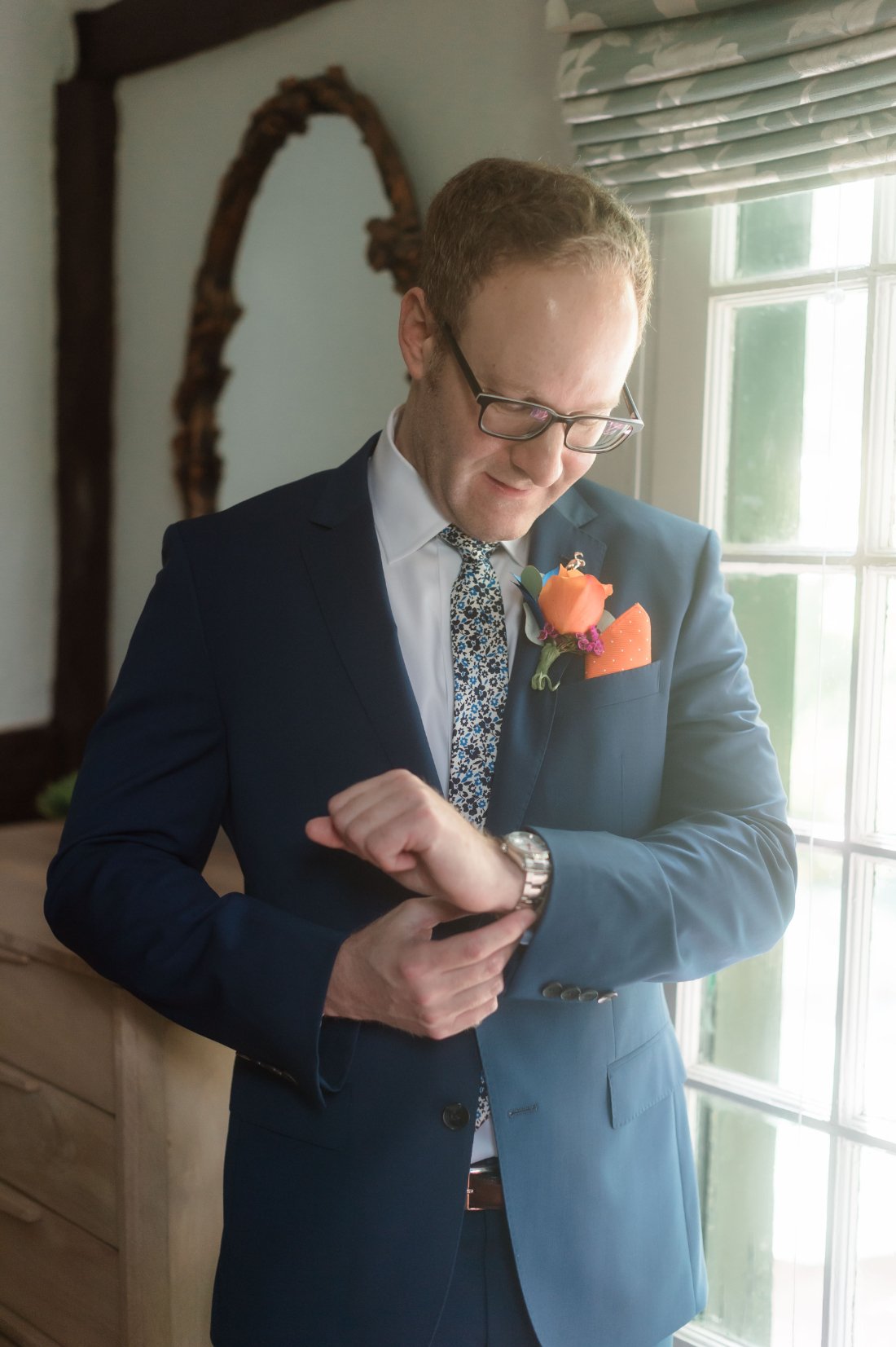   A groom in a navy suit adjusts his watch while standing near a window. He wears a floral tie, glasses, and an orange boutonniere, smiling quietly during a morning getting-ready moment.    