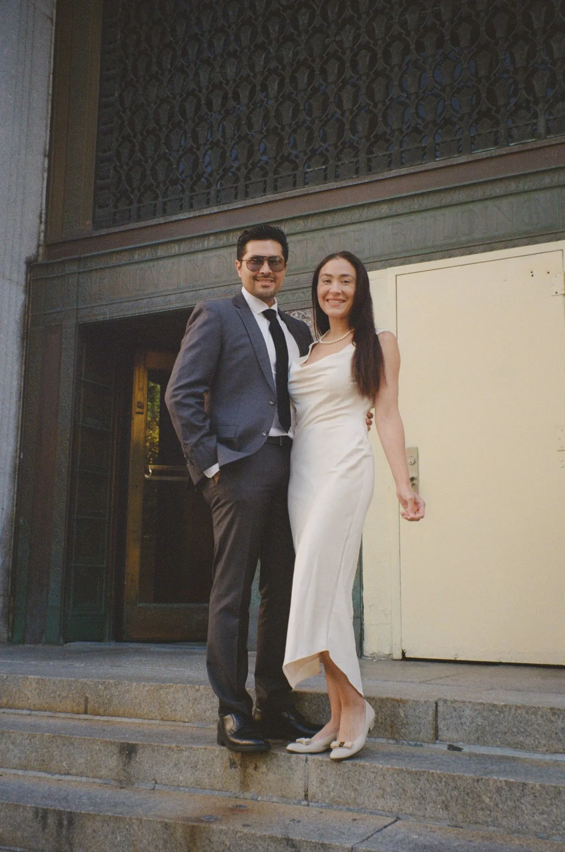   A couple poses on the courthouse steps, smiling. The bride wears a sleek ivory dress and pearls, while the groom wears a suit and tie. They stand closely together in front of a tall building.    
