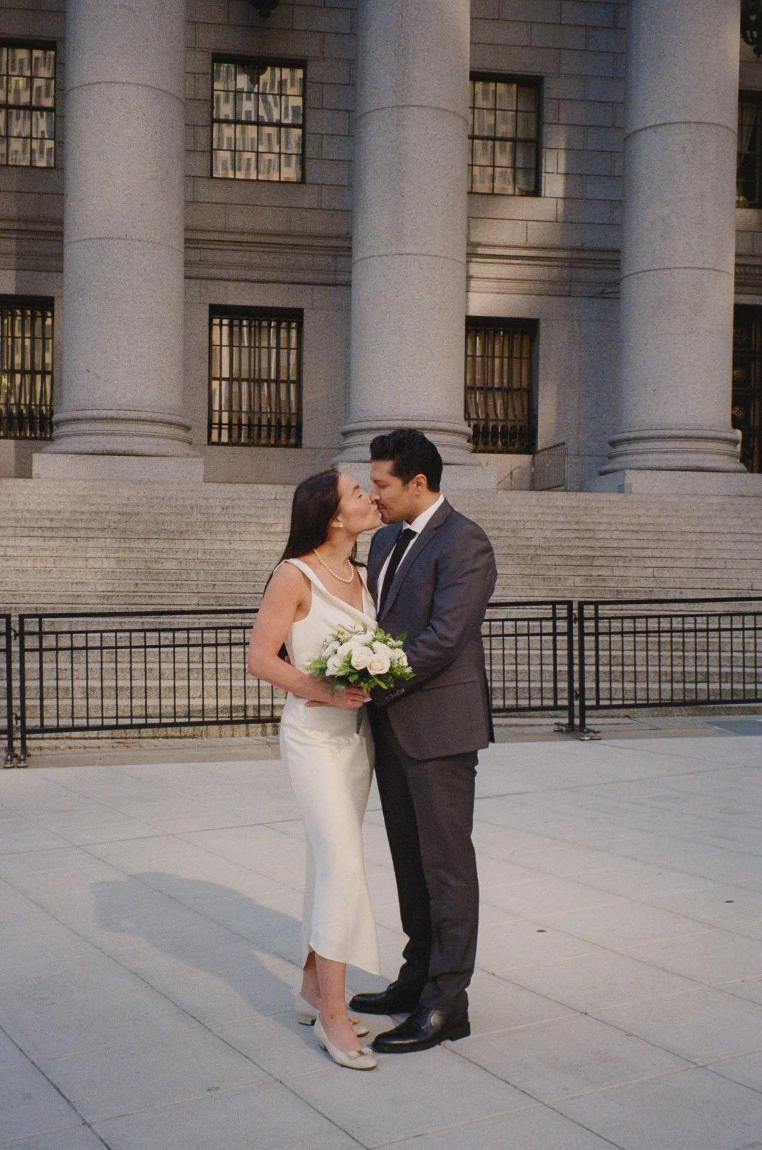   A bride and groom share a kiss outside a courthouse. The bride holds a small bouquet of white roses while the couple stands in front of large stone columns and wide steps.    