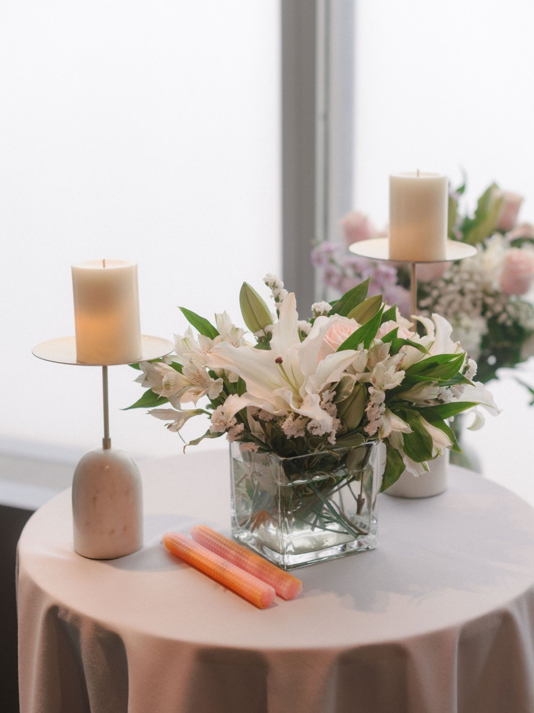   A round cocktail table with a modern floral centerpiece of white lilies and pink roses, flanked by two pillar candles and pastel taper candles, placed near a bright window at a wedding reception.    