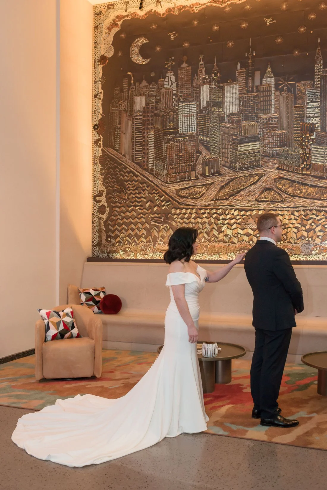   A bride gently taps her groom’s shoulder for their first look moment. They stand in front of a large, golden city mural. She wears a fitted off-shoulder gown with a long train.    